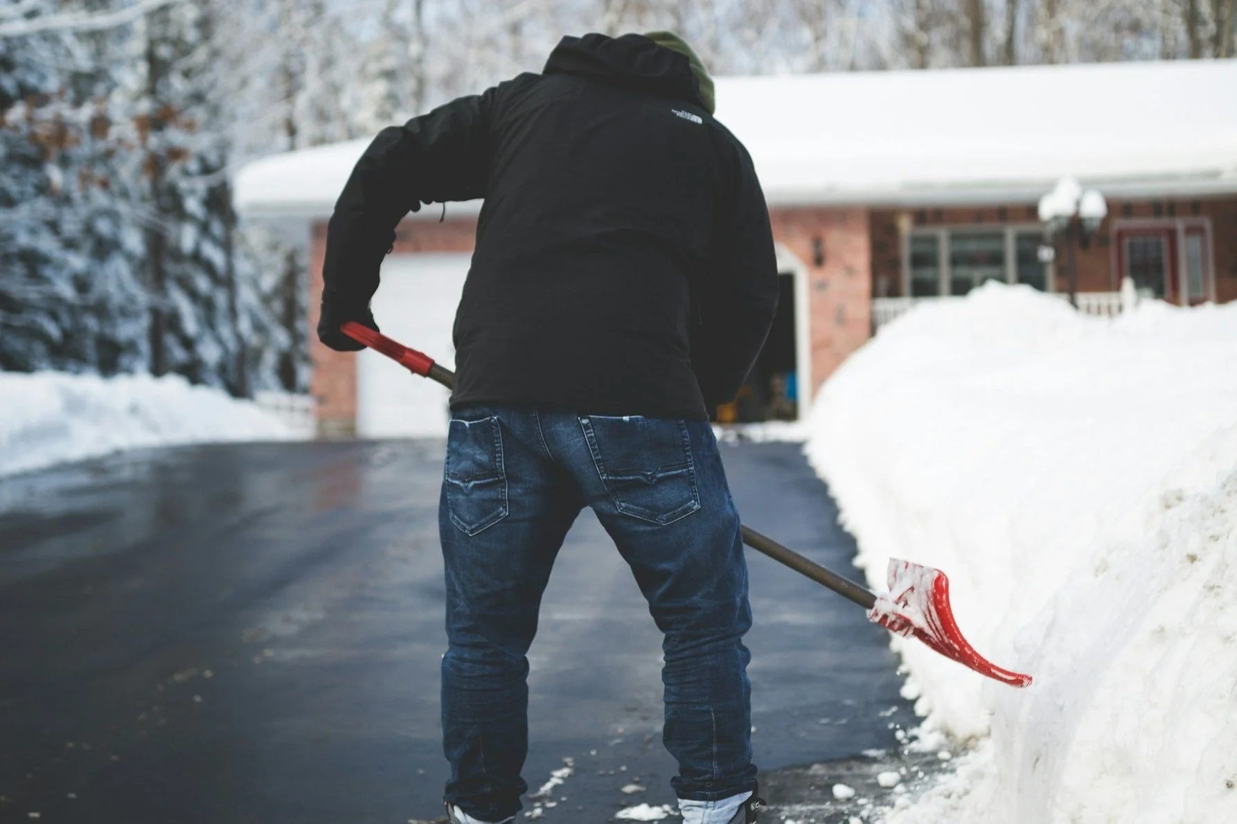 Man shoveling snow from a driveway near a house with snow-covered trees in the background.