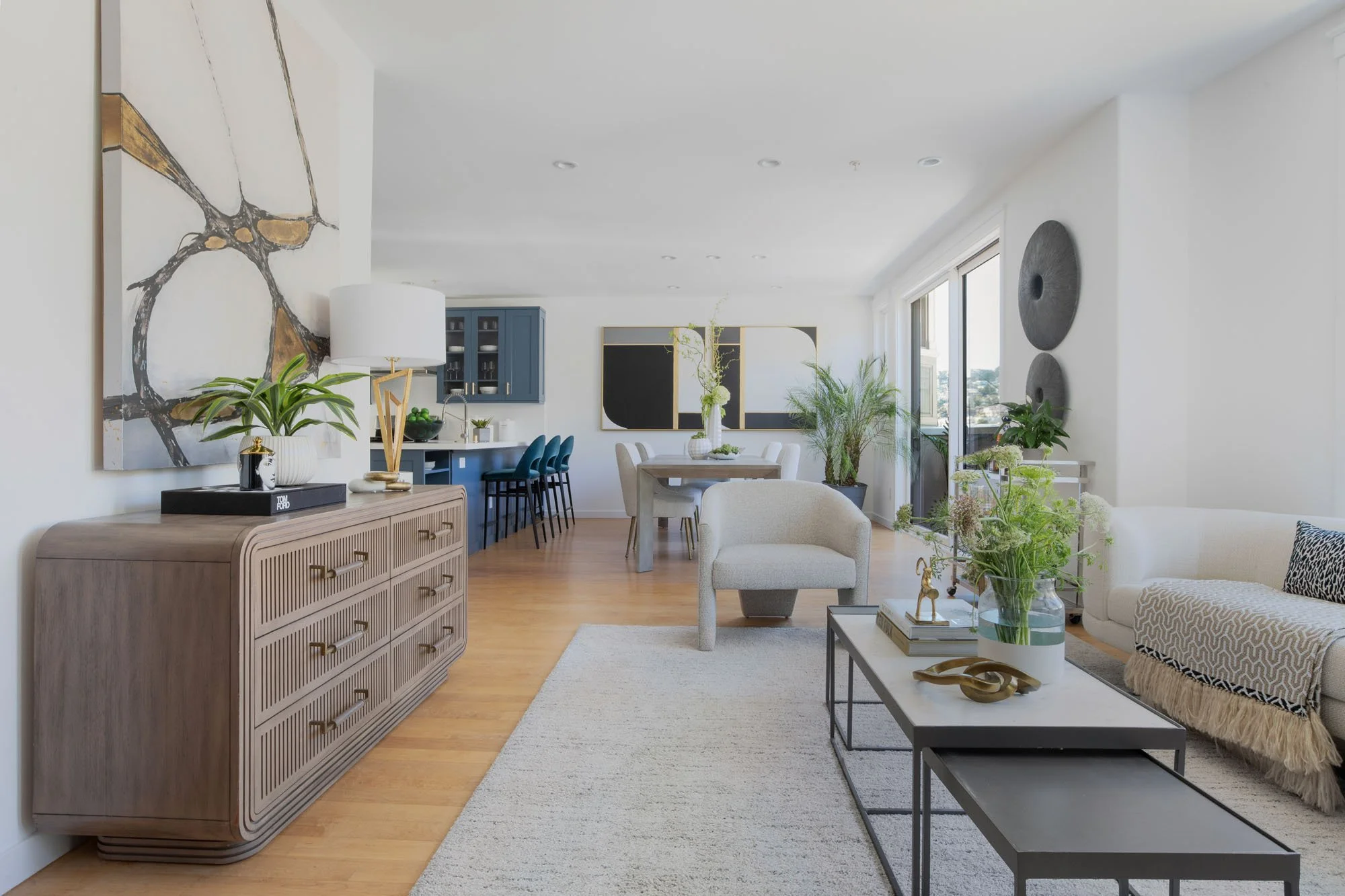 Wide-angle view of a luxury open-concept living room featuring a fluted wood dresser, abstract gold and white wall art, and a glimpse into the slate blue kitchen.