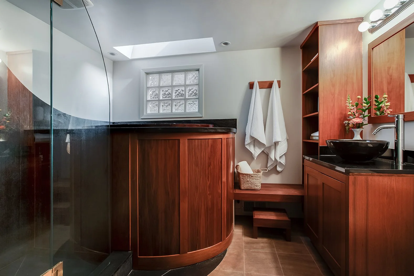 Custom spa-like primary bathroom featuring warm wood cabinetry, a black vessel sink, and natural light from a glass block window and skylight.