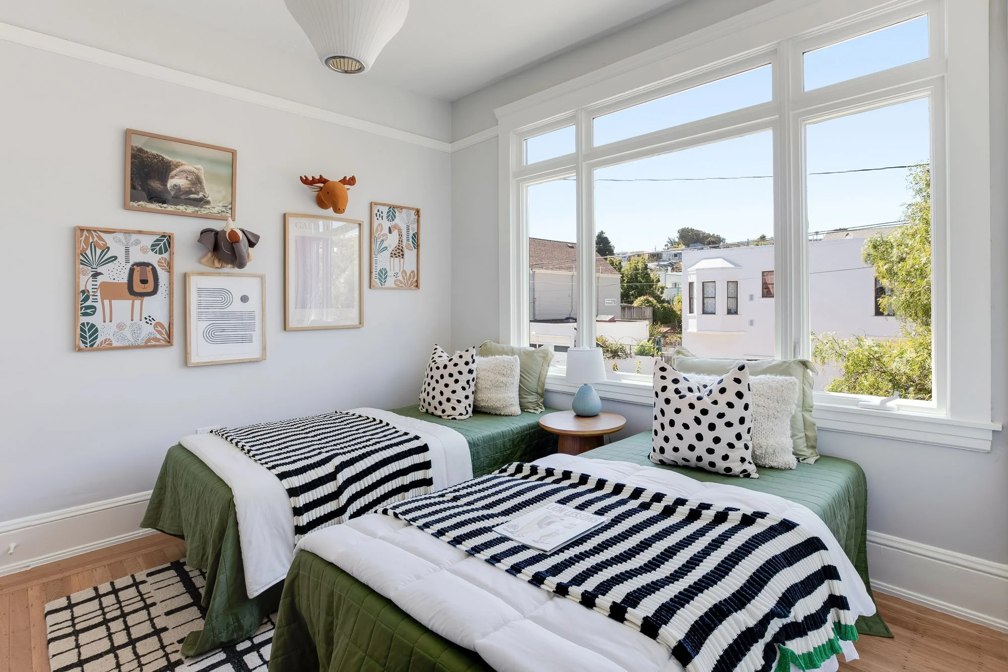 Staged children’s bedroom featuring twin beds with green linens and striped blankets, animal-themed wall art, and a large window overlooking the neighborhood.