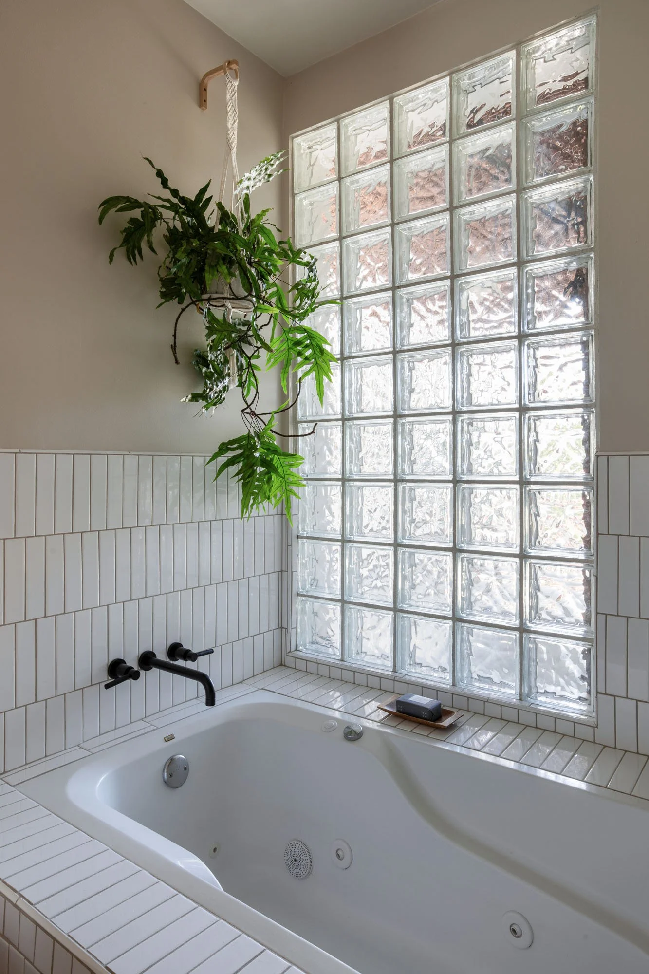 Close-up of a white whirlpool soaking tub with matte black fixtures, a glass block window for natural light, and a hanging macramé plant.
