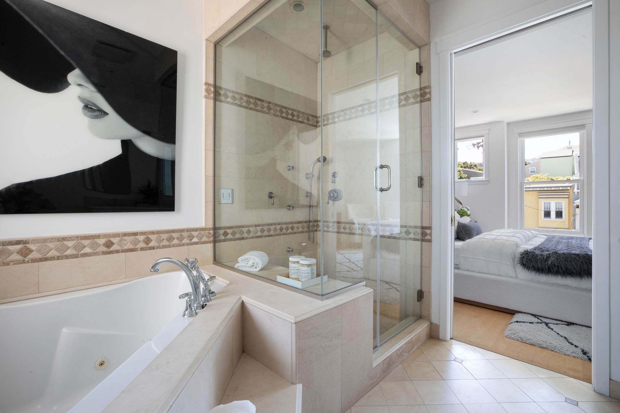 Modern guest bathroom featuring a white vanity with gold hardware, a round wood-framed mirror, and a neutral tile bathtub surround with decorative mosaic accents.