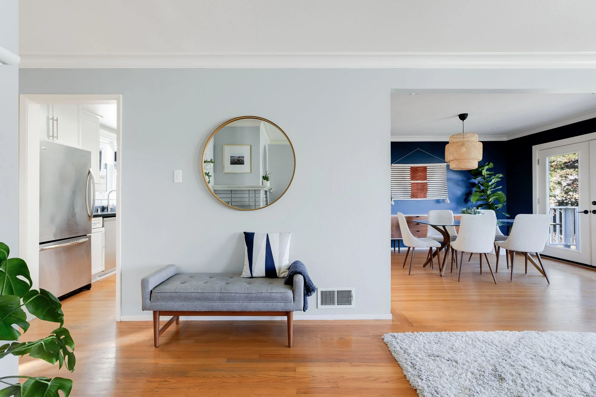 A minimalist entryway featuring a light gray wall, a round gold-framed mirror, and a gray upholstered bench with a navy blue accent pillow.