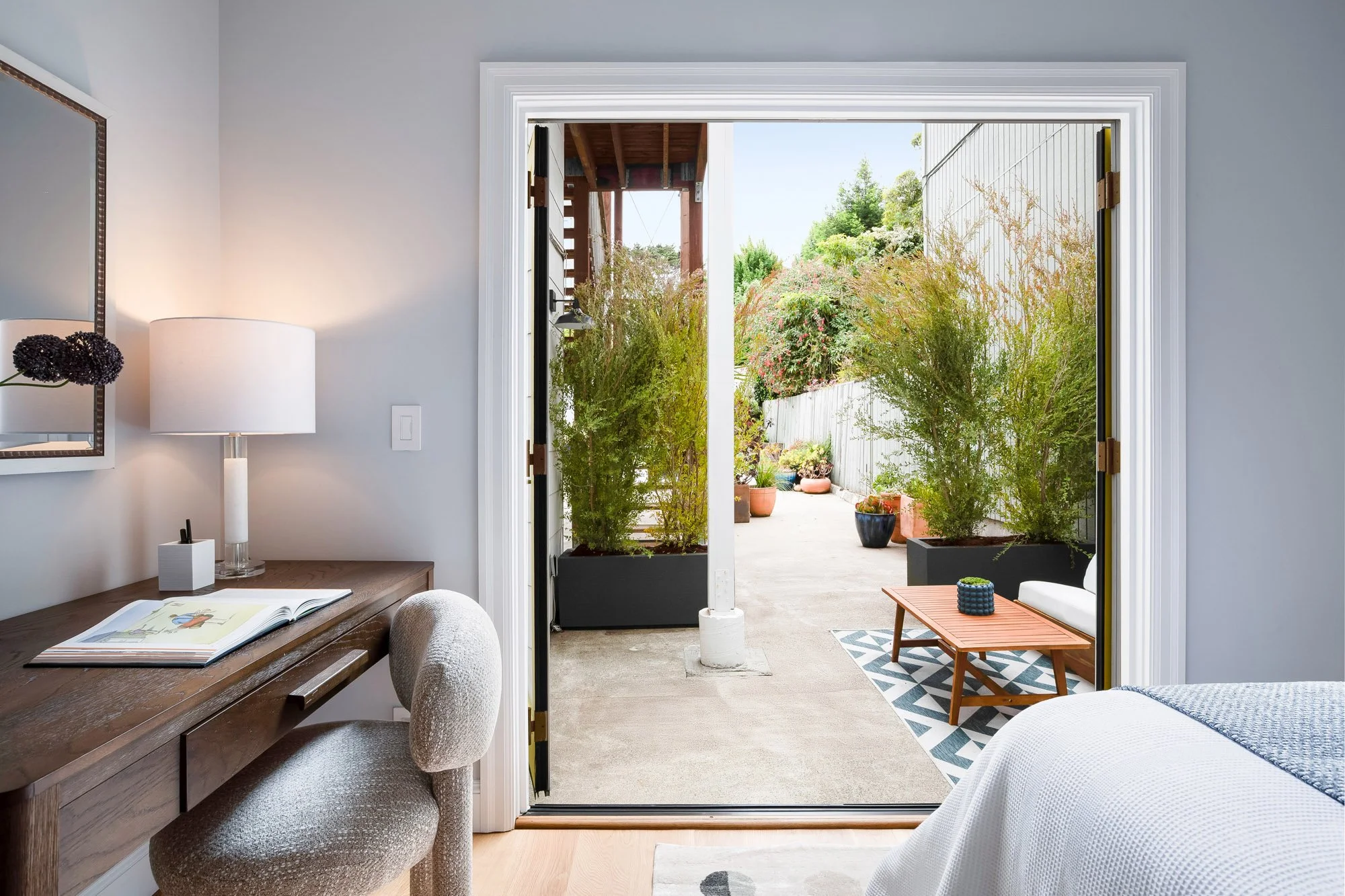 View from second bedroom through open bifold glass doors to private ground-level patio at 4545 18th Street San Francisco TIC, with walnut desk, tall ornamental grasses in black planters, and outdoor seating area beyond