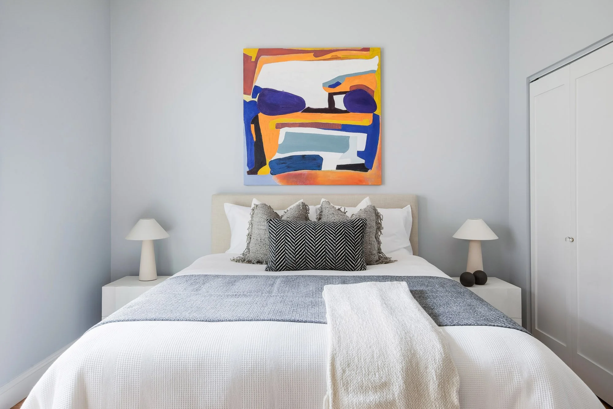 Second bedroom detail at Castro San Francisco luxury TIC featuring upholstered platform bed with layered gray and white bedding, bold orange and blue abstract artwork, matching white cone table lamps, and built-in closet