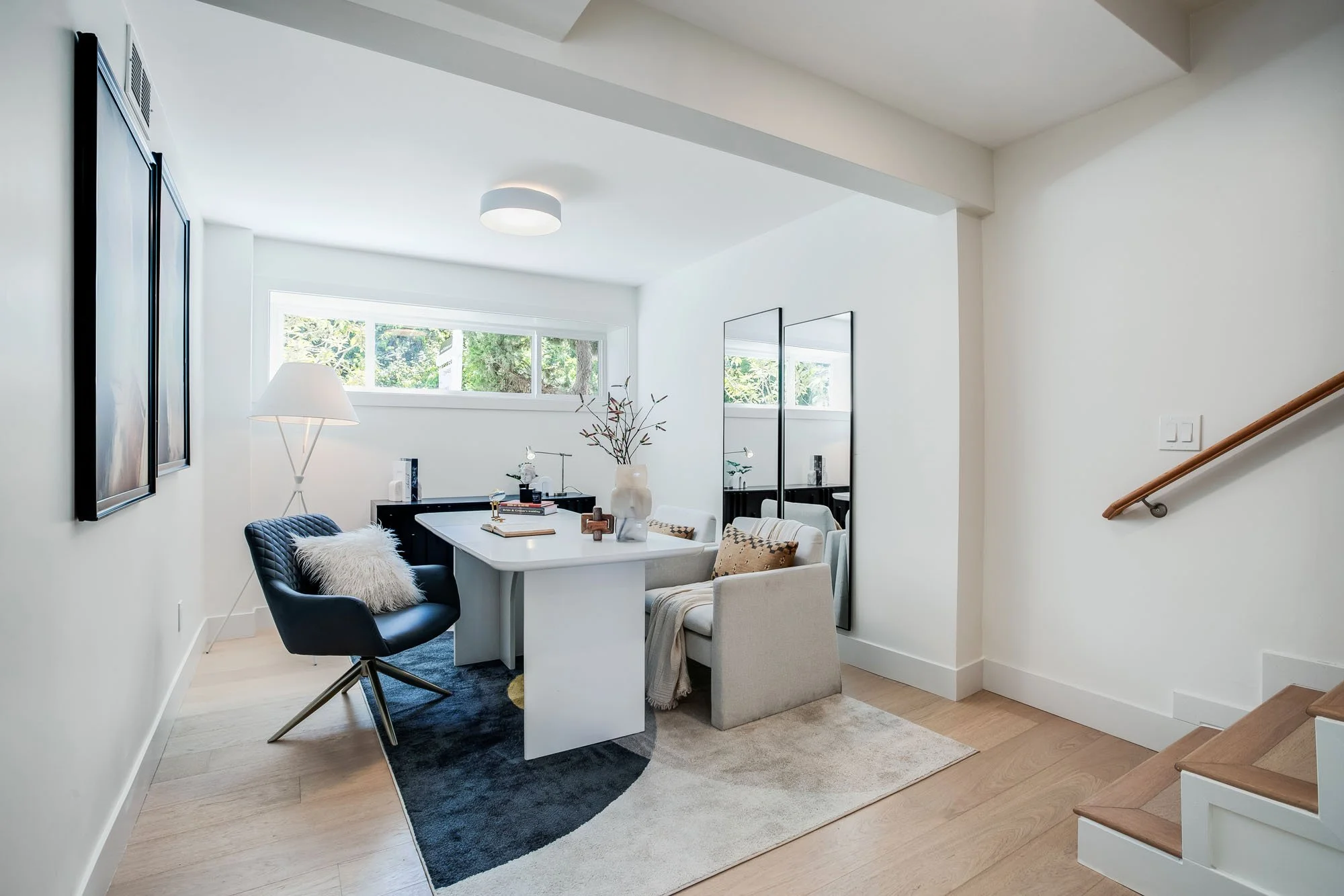 A modern home office with white walls, light wood floors, and a sleek white desk. A navy blue swivel chair features a plush white pillow, set atop a geometric blue and grey area rug. Large floor-to-ceiling mirrors and a wide window overlooking greene