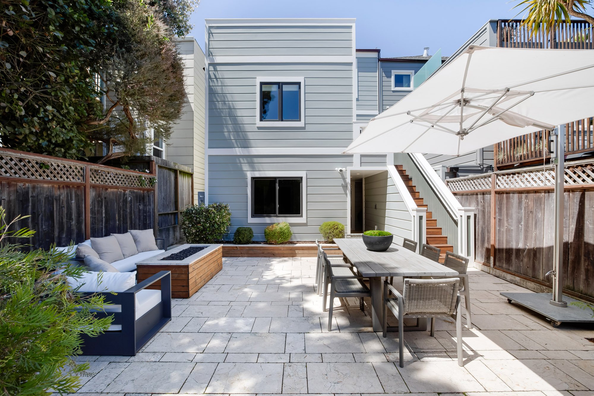 Rear elevation of 771 Elizabeth Street in Noe Valley showing gray siding, the staircase down to a flat paver patio, lounge seating, and a built-in planter beneath sunny open sky.