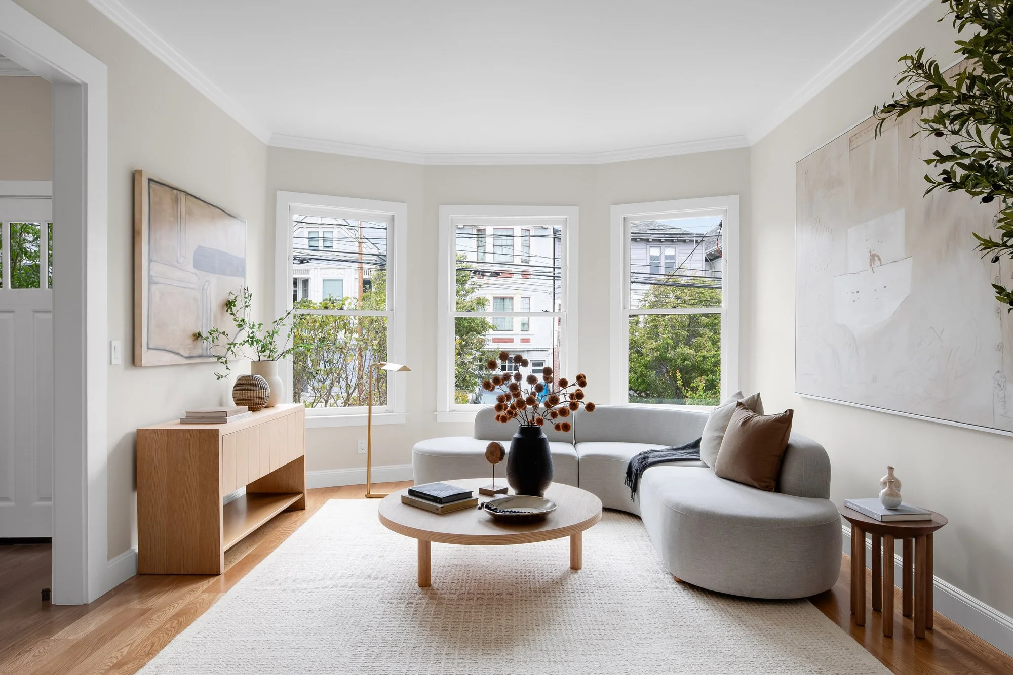 Light-filled living room at 771 Elizabeth Street in Noe Valley with a classic Edwardian bay window, curved sofa, round oak coffee table, oversized art, and hardwood floors.