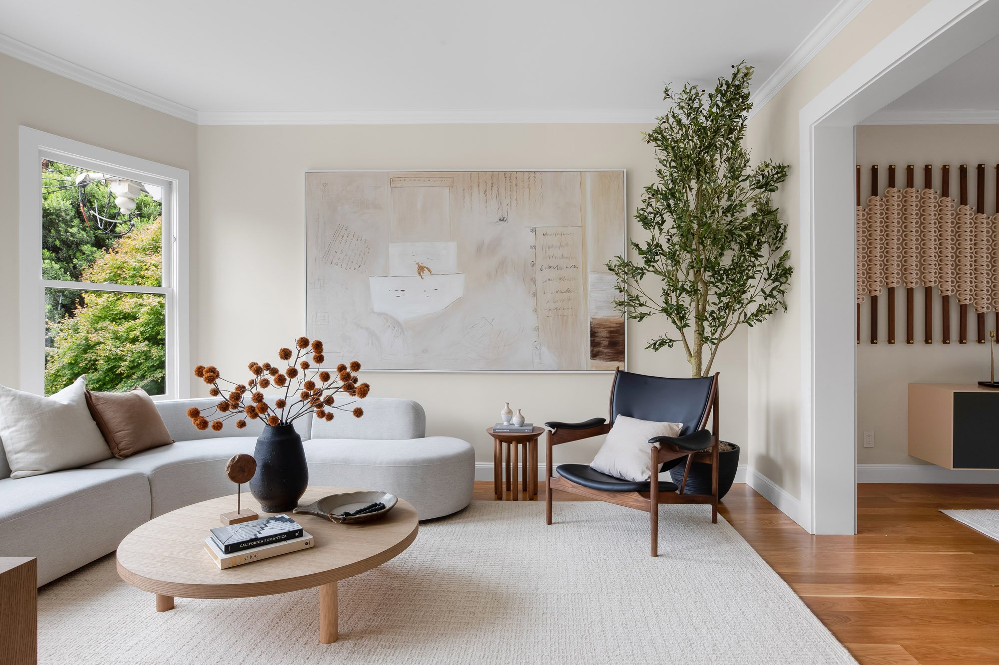 Living room at 771 Elizabeth Street featuring a curved sofa, leather-and-walnut lounge chair, olive tree, and large-scale artwork, with an open view through to the dining room's Windy Chien installation.