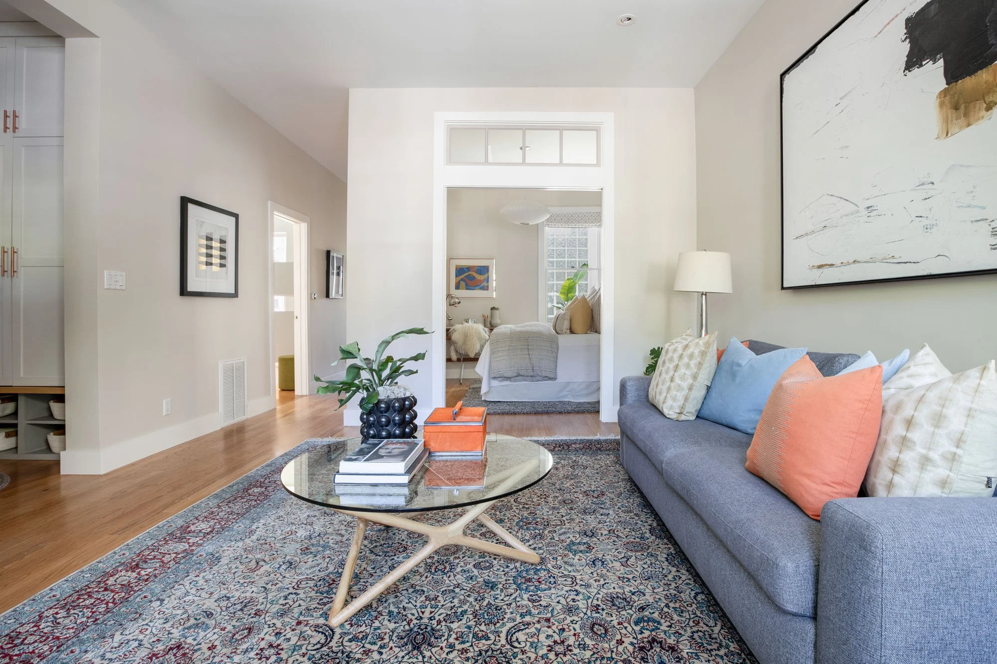 Open-concept living space showing a grey sofa and glass table leading into a bright bedroom through a white doorway with a glass transom window.