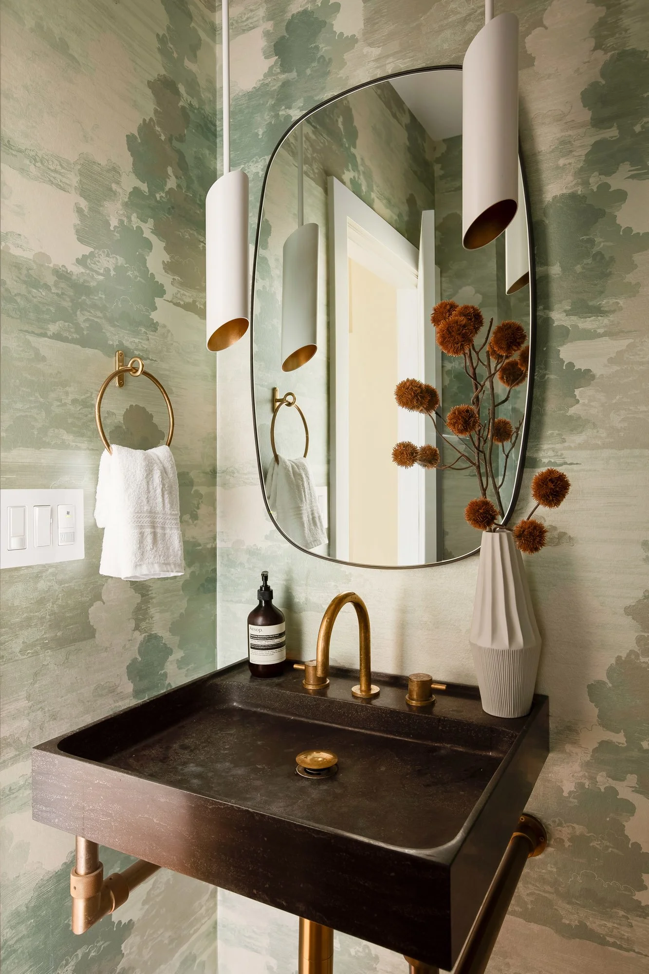 Designer powder room at 771 Elizabeth Street featuring cloud-motif wallpaper, a honed stone trough sink, brass fixtures, cylindrical pendant lighting, and a curved black-framed mirror. 