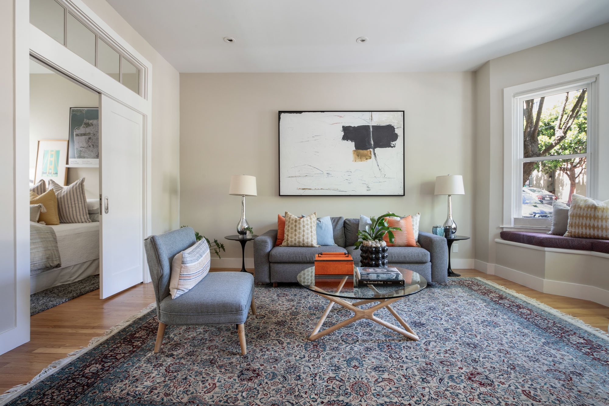 Modern living room with a large abstract painting, oriental area rug, glass coffee table, and a built-in bay window seat with street views.