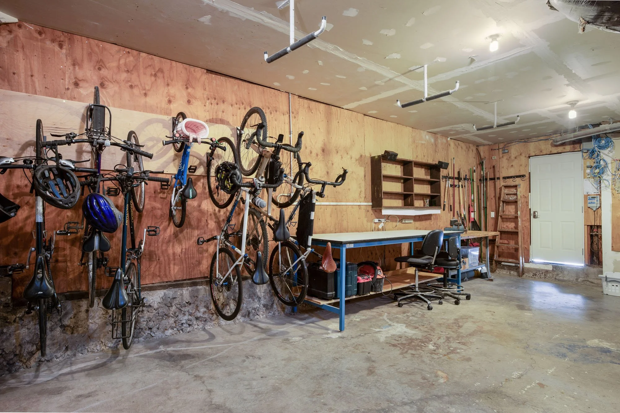 Organized garage storage area featuring multiple bicycles hanging on a plywood wall, a long blue metal work table, and overhead storage racks.