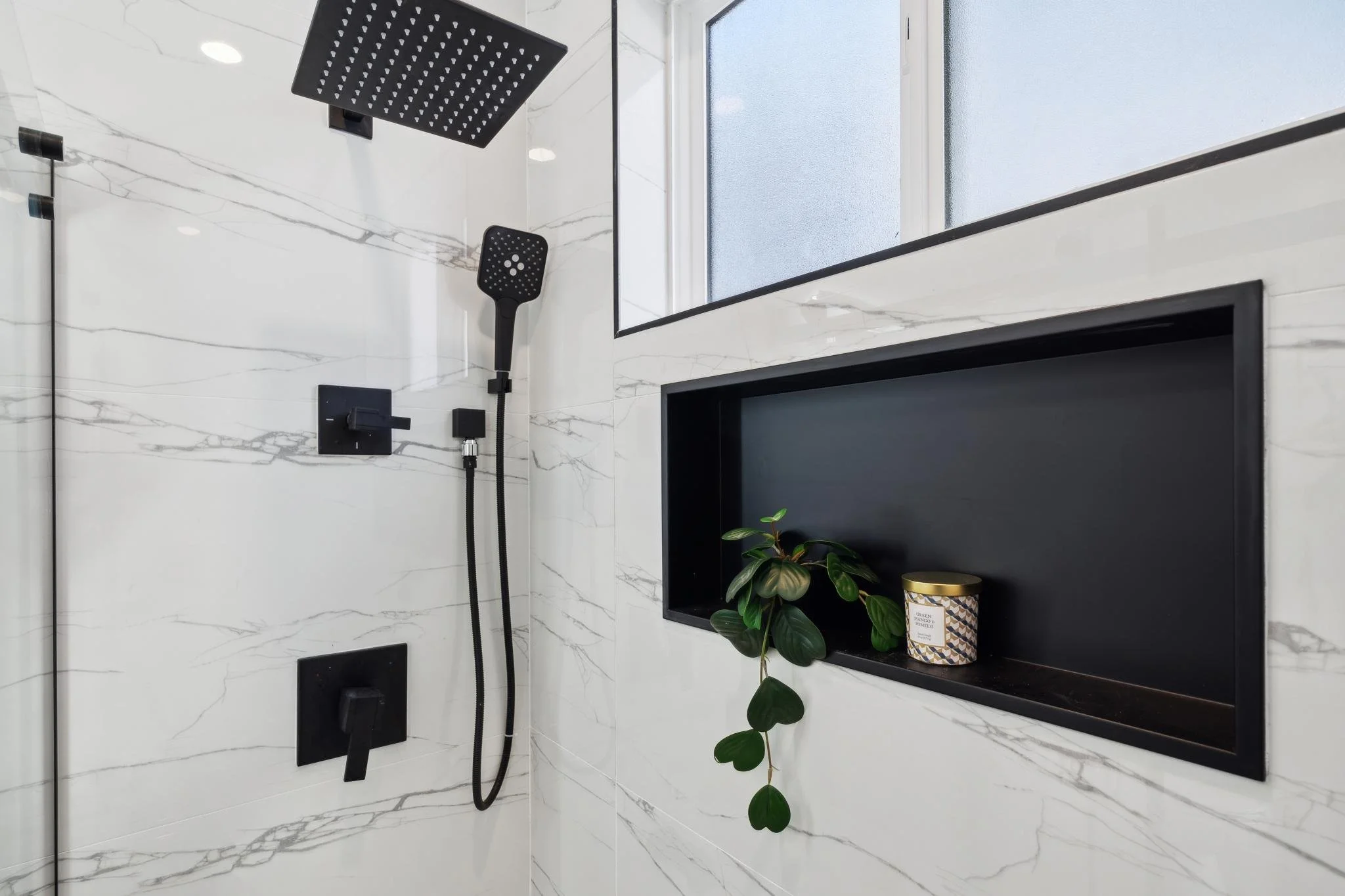 Detailed view of a luxury walk-in shower featuring a matte black rain showerhead, matching hand wand, and a recessed black storage niche with Calacatta marble surrounds.
