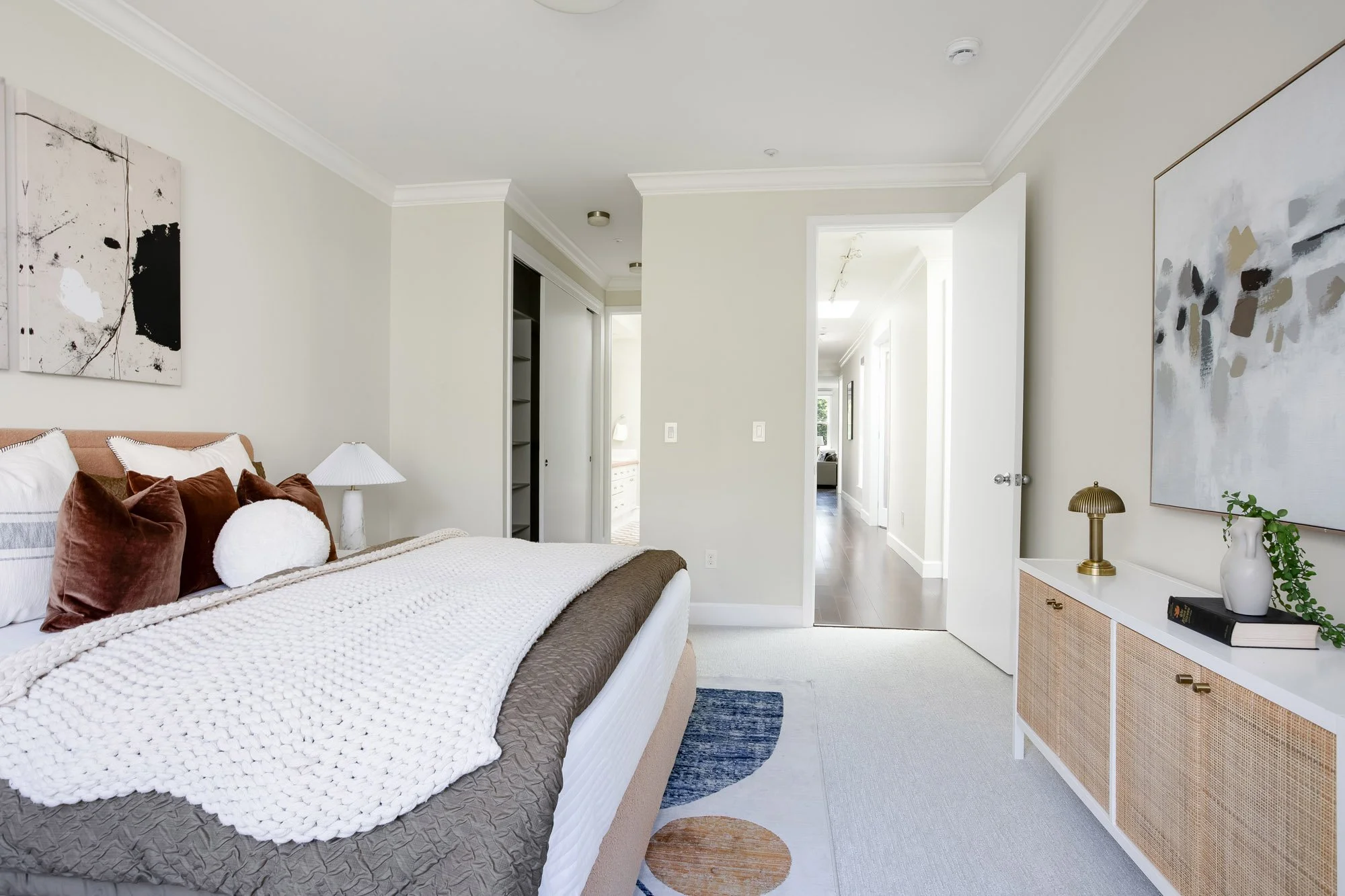 Primary bedroom at 2172 Fell Street San Francisco with blush upholstered king bed, rattan and white credenza, brass mushroom lamp, built-in closet, crown molding, and hallway view toward main living areas
