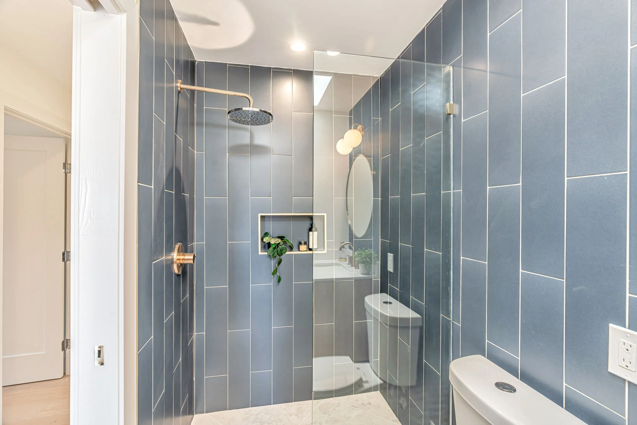 A detail shot of the walk-in shower in the hallway bathroom, showing the striking blue-grey vertical tiles and a high-end gold rain showerhead. A built-in wall niche holds bath products and a small plant.