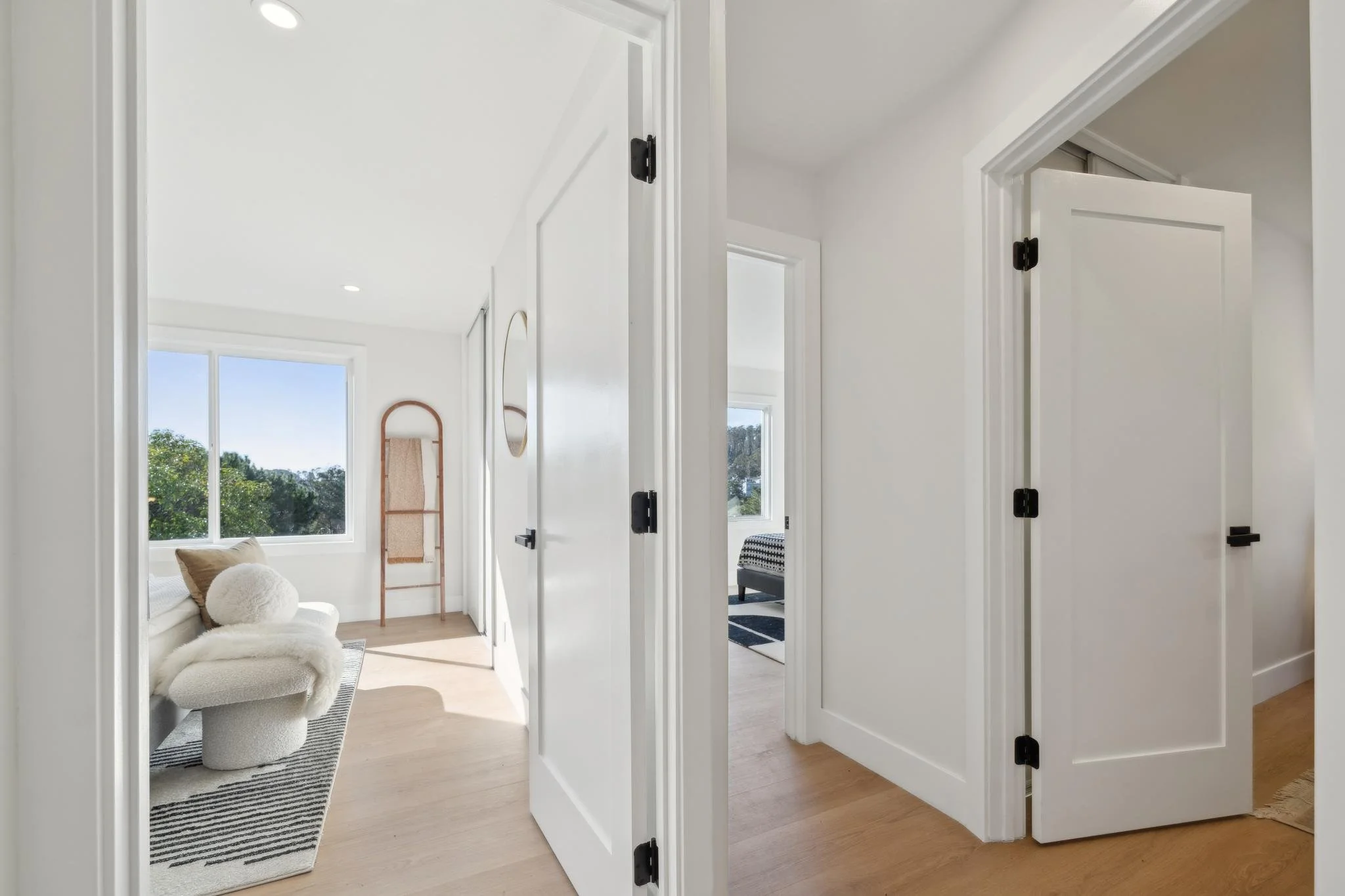 Bright hallway leading into a sun-drenched primary bedroom with light hardwood floors and minimalist white doors featuring matte black hardware.