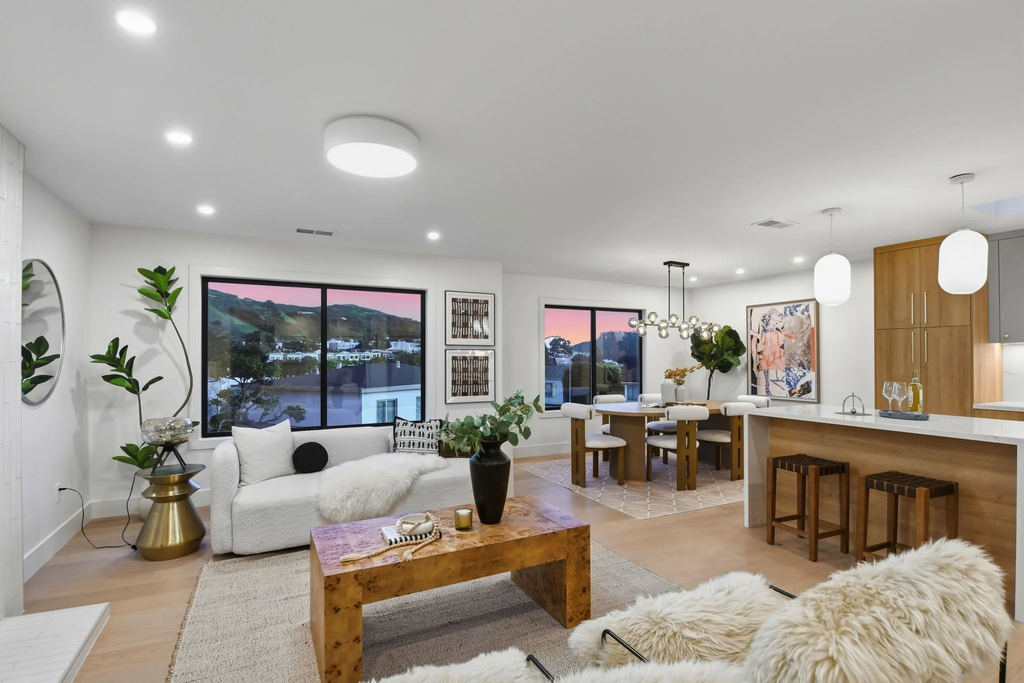 Luxury open-concept great room at sunset, featuring a waterfall island, designer furniture, and panoramic views of the San Francisco hills under a pink sky.