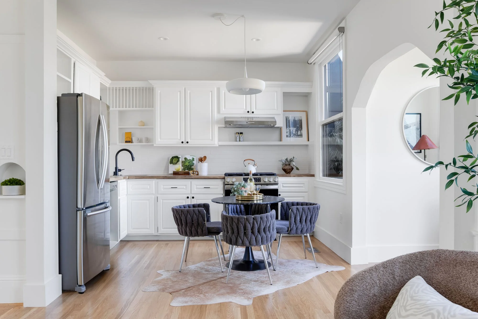 Modern white kitchen with stainless steel appliances, butcher block countertops, and a dining nook featuring chic woven chairs and a cowhide rug.