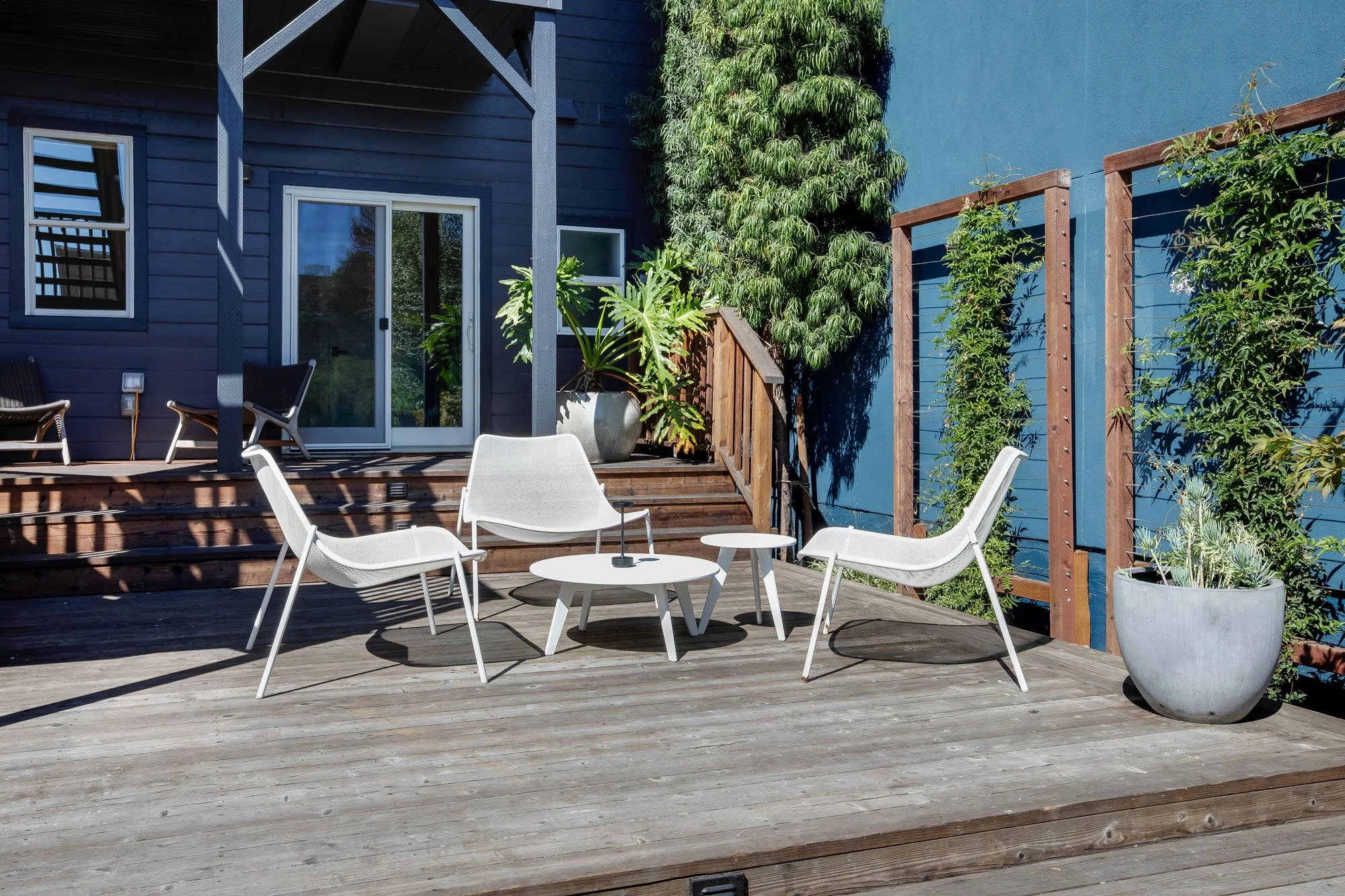A spacious, multi-level wooden entertaining deck at 246 Detroit featuring modern white lounge chairs and a matching coffee table, accented by lush garden greenery and a blue accent wall.