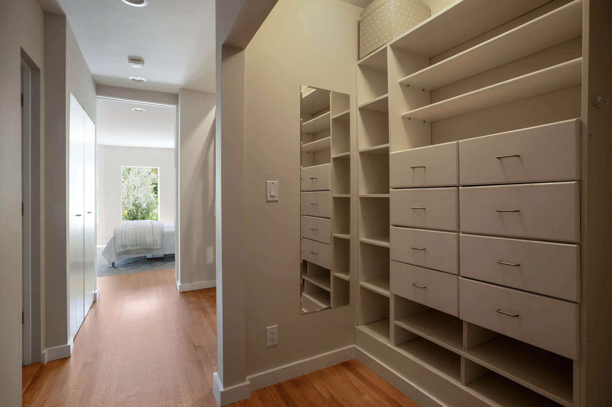 Custom walk-in closet with built-in white shelving, multiple drawers, a full-length mirror, and hardwood floors leading toward a bright bedroom.