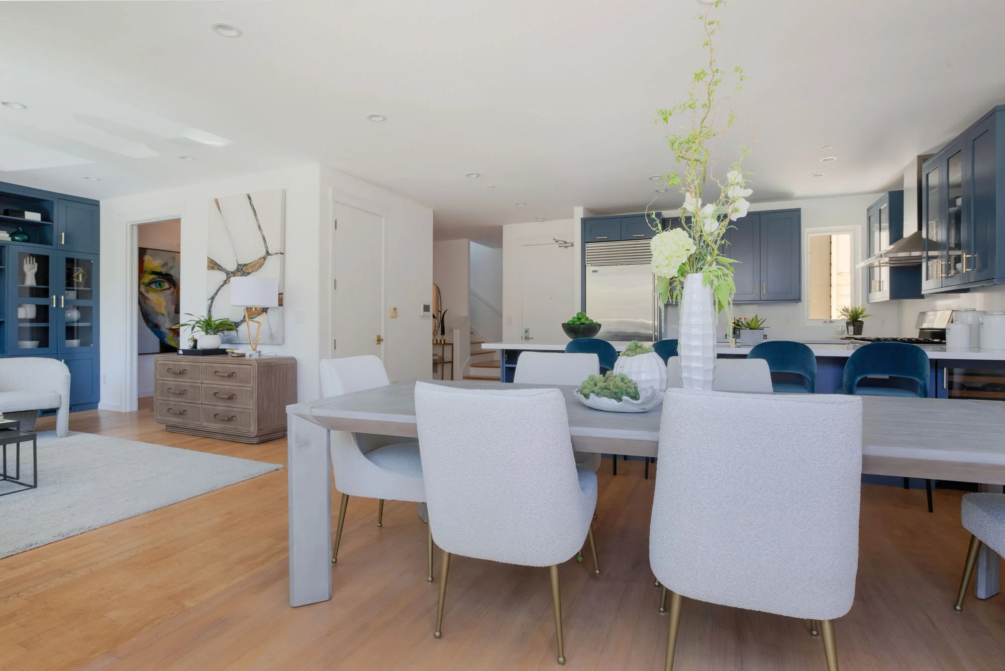 Open-concept living and dining room featuring a light grey wood dining table, white upholstered chairs, and a view into a kitchen with navy blue shaker cabinets.