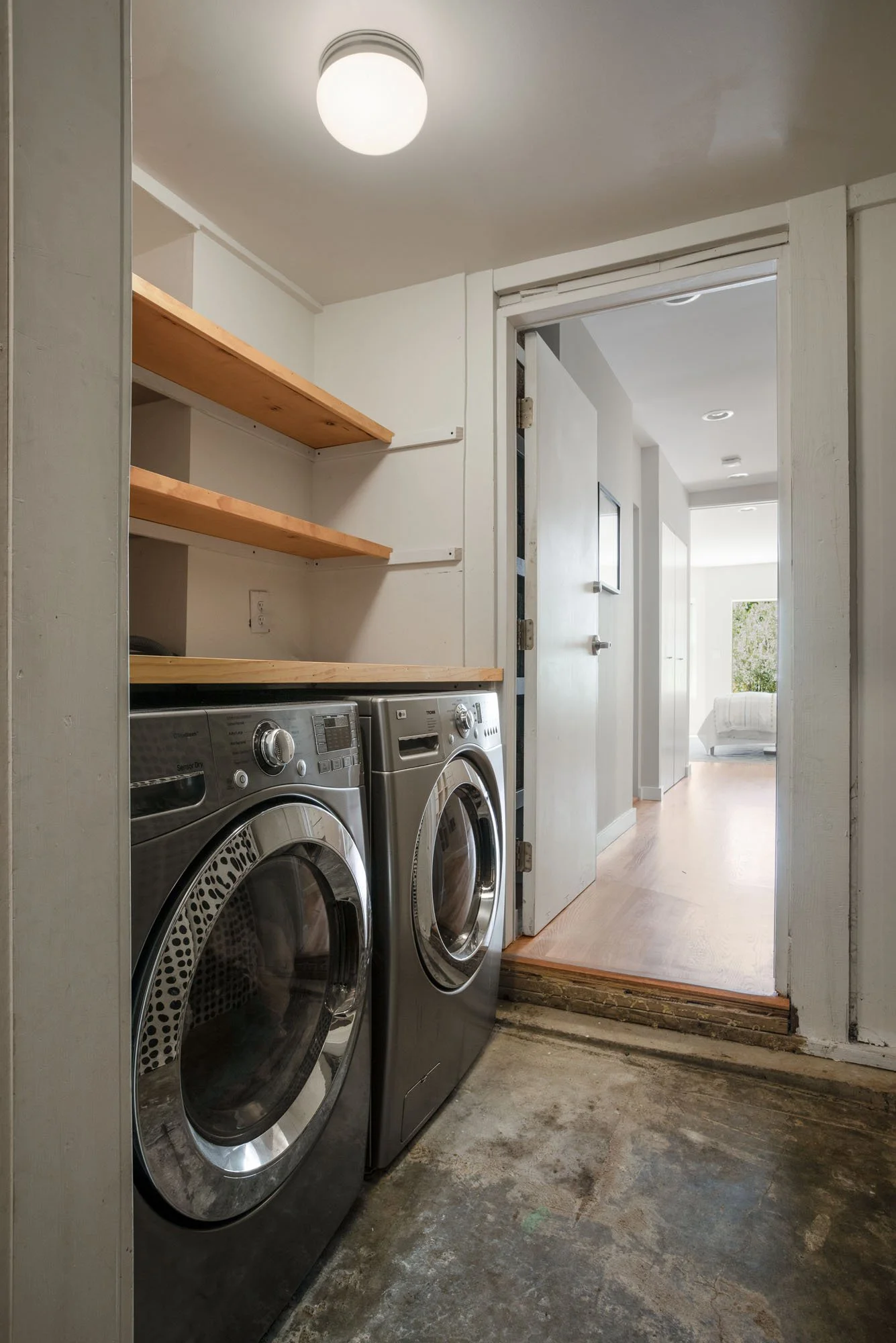 Functional laundry room with front-loading stainless steel washer and dryer units, natural wood shelving, and a doorway leading to the main hallway.