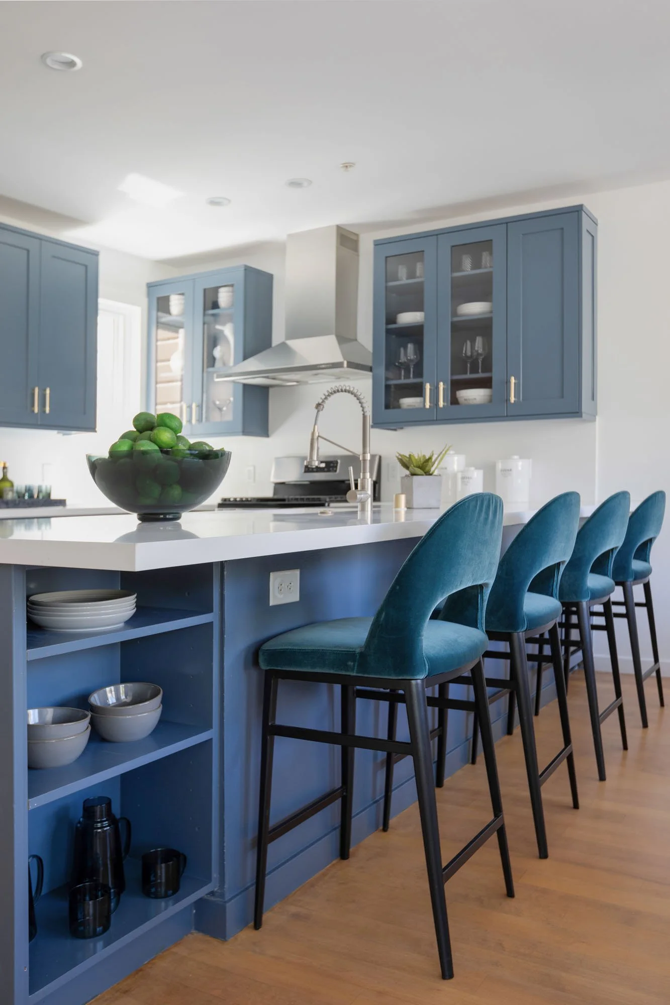 A side view of the kitchen island highlighting a row of four teal velvet barstools with black legs. The end of the island includes built-in open shelving holding grey bowls and black mugs, while the countertop holds a large glass bowl of limes.