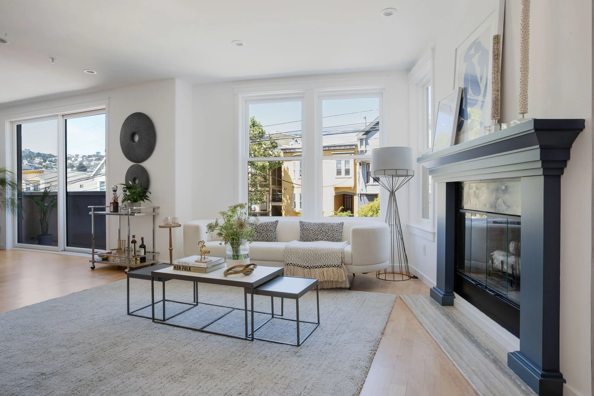 A bright living room featuring a white curved sofa accented by patterned pillows and a throw blanket. A blue-painted fireplace mantle serves as a focal point, with a large grey area rug and a multi-tiered black coffee table in the center of the room.