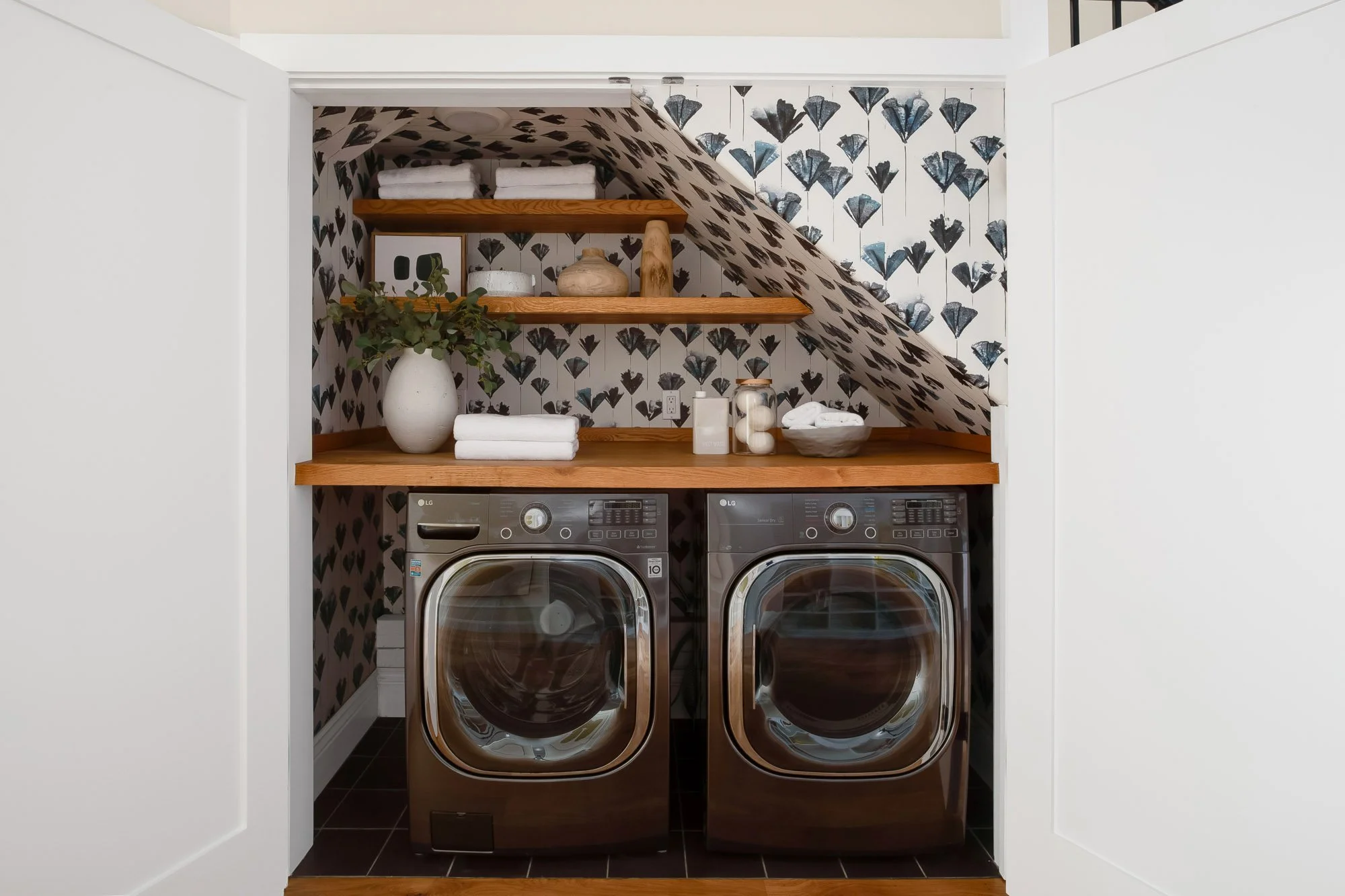 Finished laundry room at 771 Elizabeth Street in Noe Valley with a side-by-side LG washer and dryer, butcher block counter, open oak shelving, and botanical wallpaper tucked beneath the stair. 