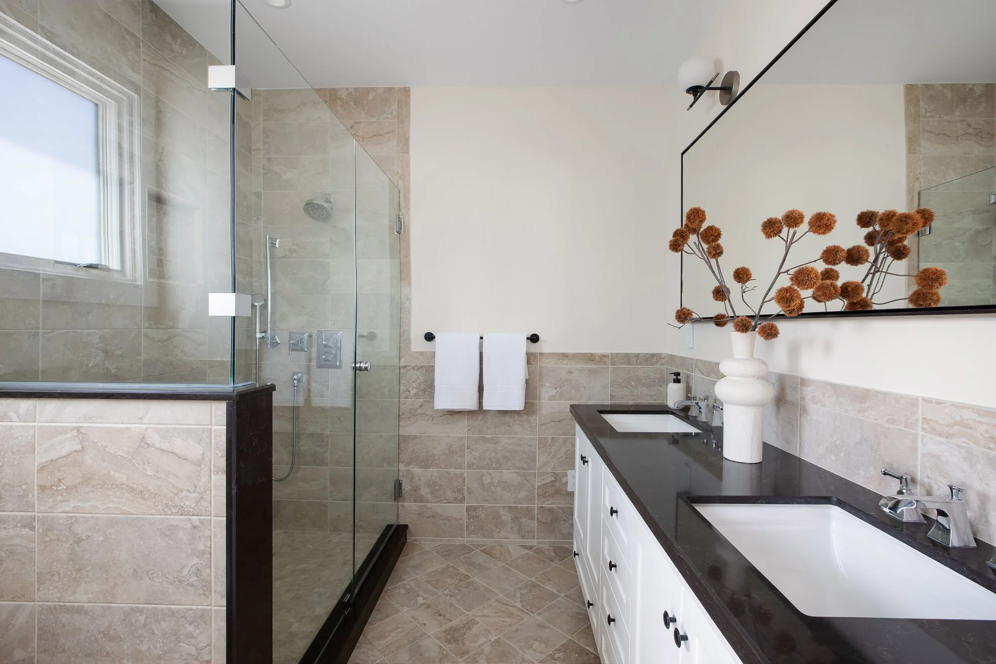 Primary en-suite bathroom at 771 Elizabeth Street with a glass-enclosed travertine shower, dual-sink vanity, dark stone countertops, and a window bringing in natural light.