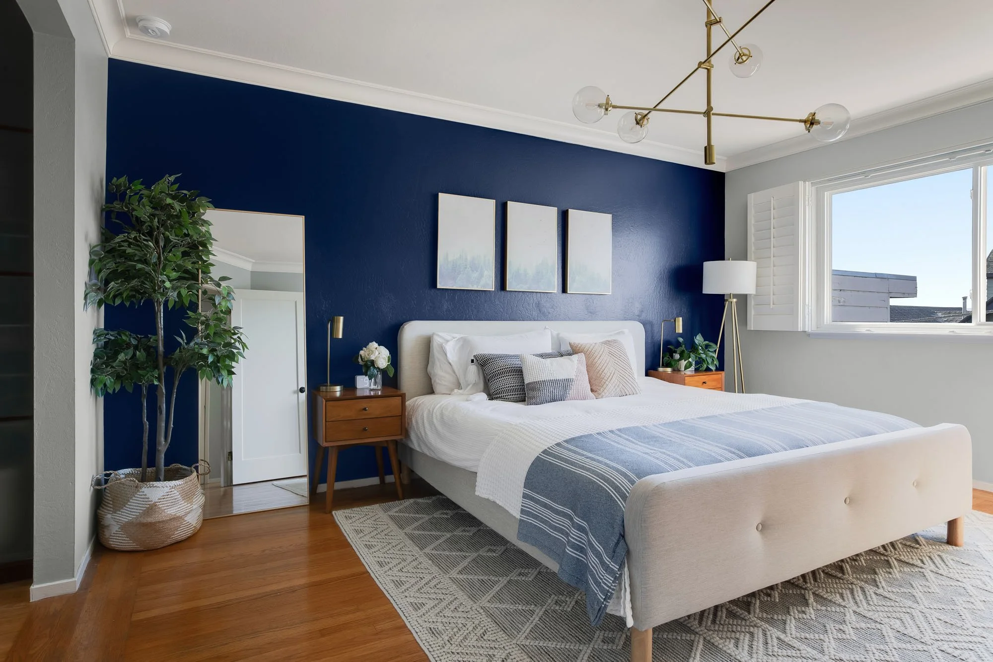 Close-up of a staged king bed with blue and white linens against a navy blue wall with triptych forest art and a full-length leaning mirror.