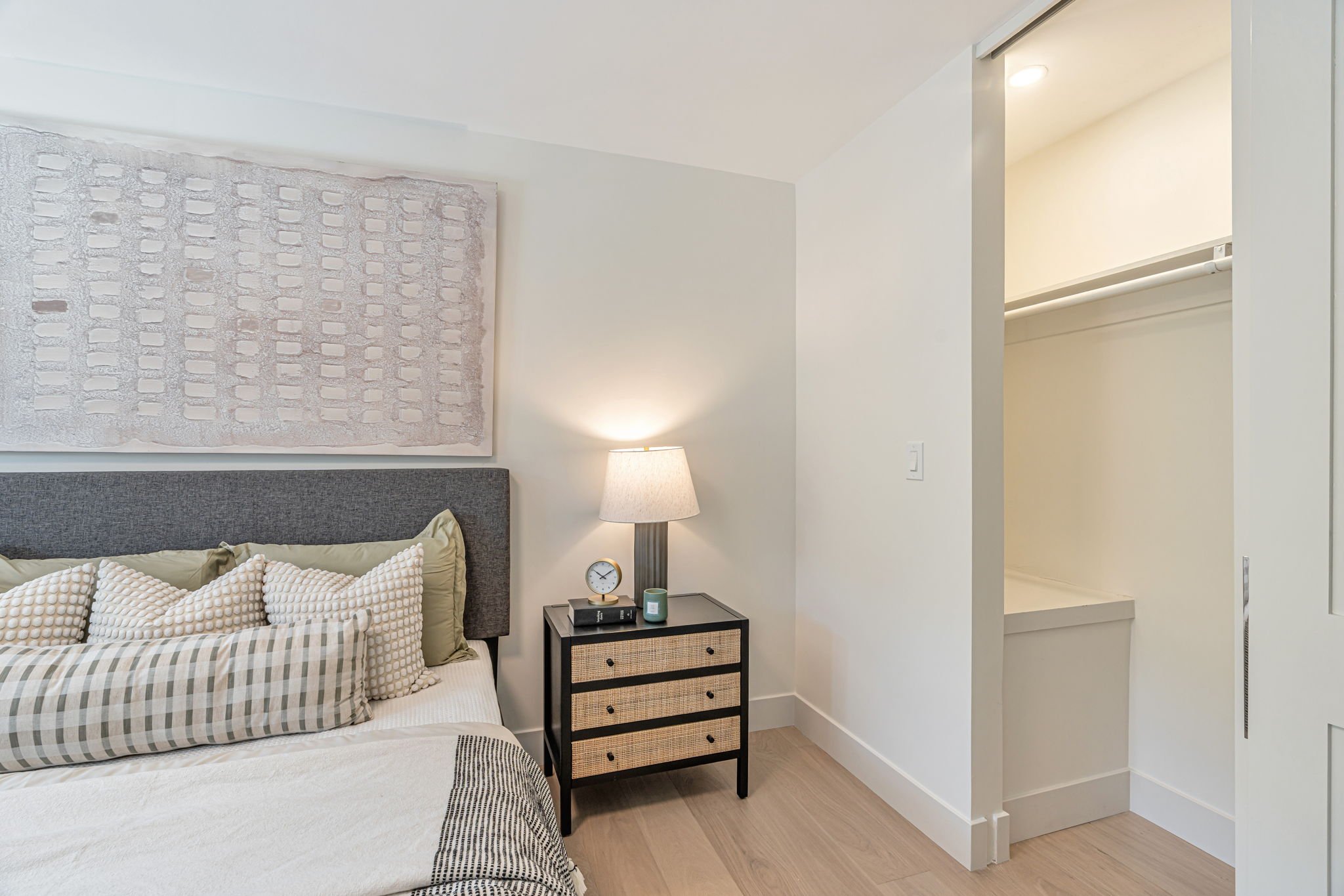 A close-up of the bedside area, highlighting a black and cane nightstand styled with a modern lamp and clock. To the right, a sliding door is open to reveal a bright, organized closet space.