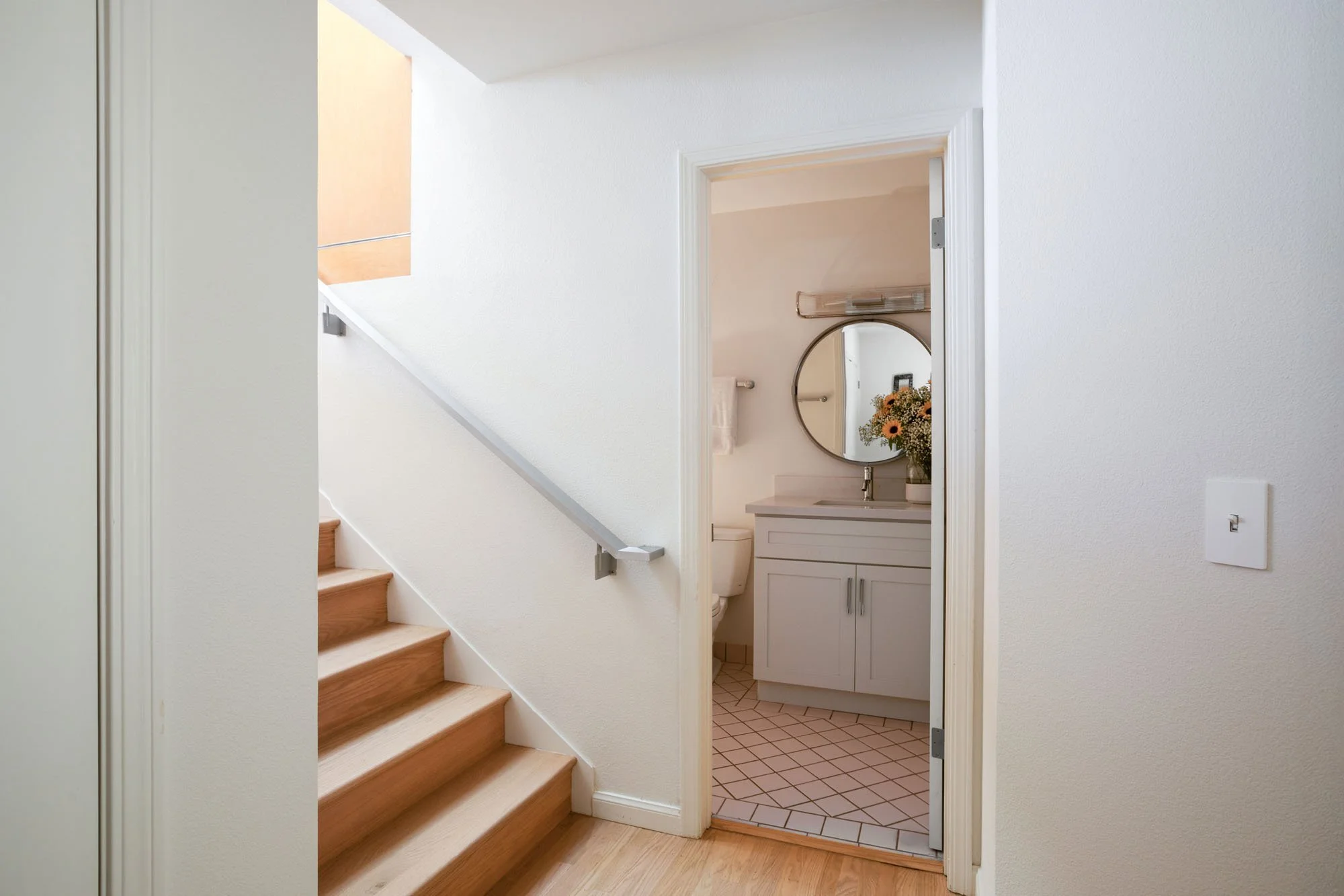 Architectural shot of a light wood staircase with a minimalist grey handrail, leading past a renovated guest bathroom with a round mirror and pinkish-toned floor tiles.