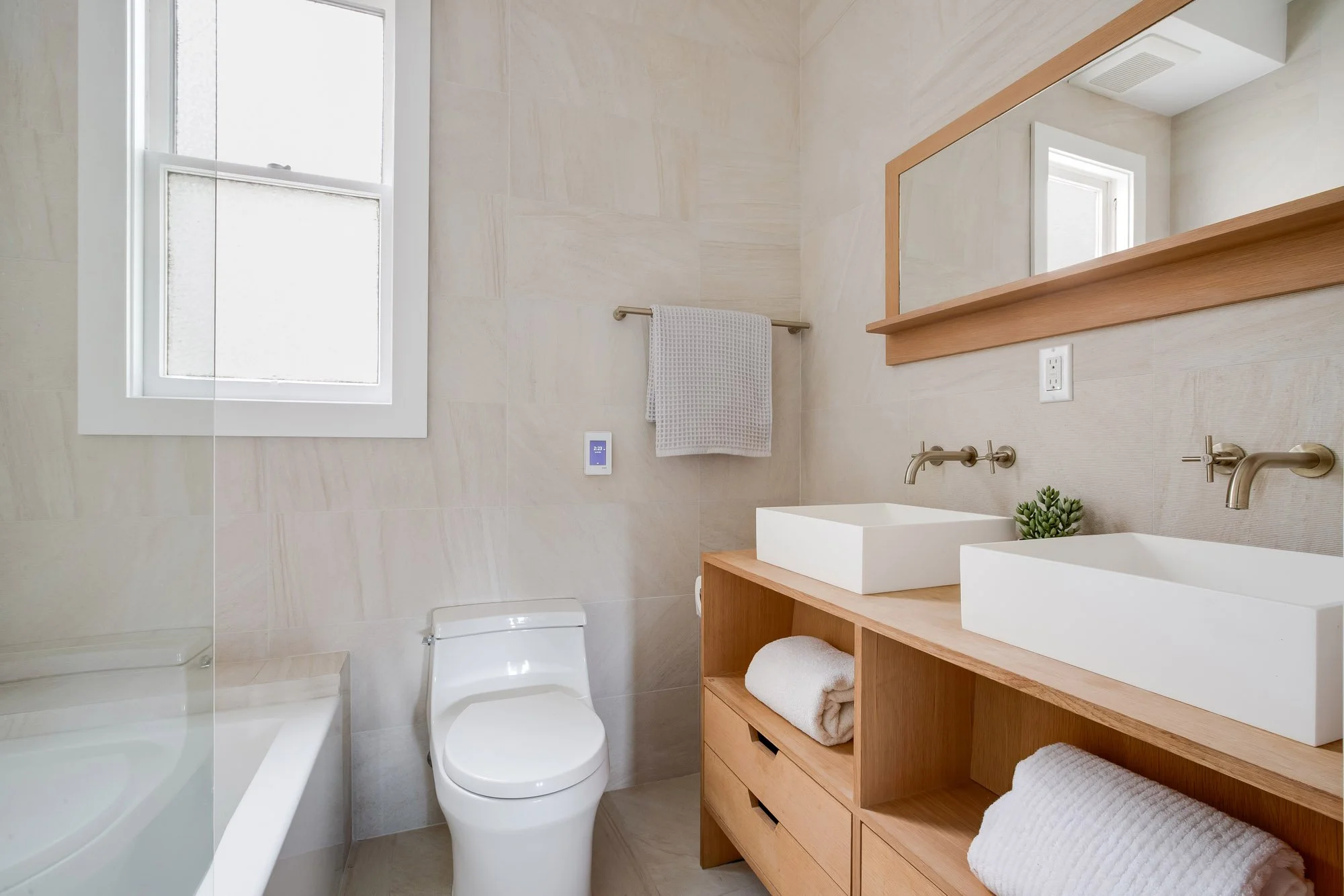 New ensuite bathroom with double white oak vanity, dual vessel sinks, wall-mount brushed nickel faucets, soaking tub, and large format stone tile in luxury Presidio Heights Victorian condo, San Francisco