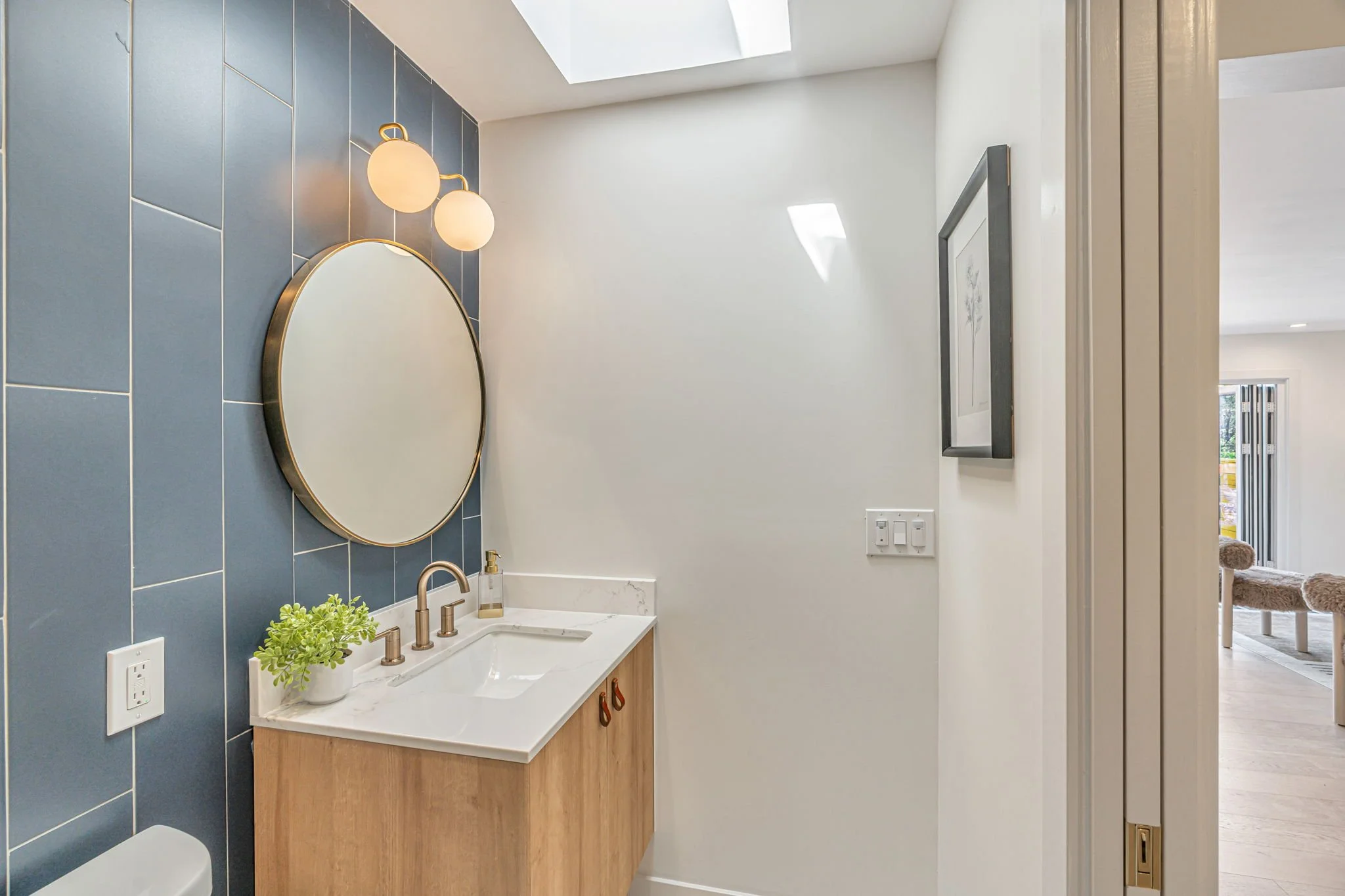A stylish bathroom featuring deep blue-grey vertical tiles and a round gold-framed mirror. A modern wood vanity is topped with white marble, and a skylight overhead provides bright, natural illumination.