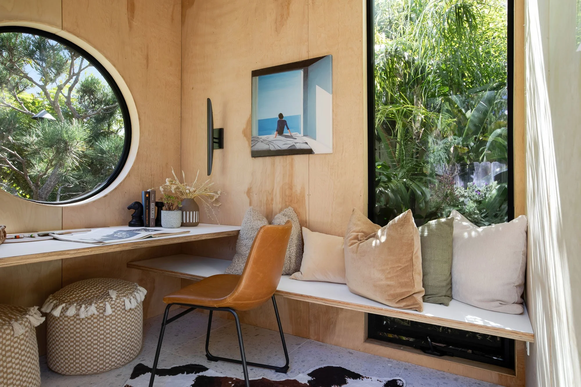 A functional home office setup inside the garden studio at 246 Detroit, featuring a plywood desk, a circular porthole window, a leather chair, and a mounted monitor.