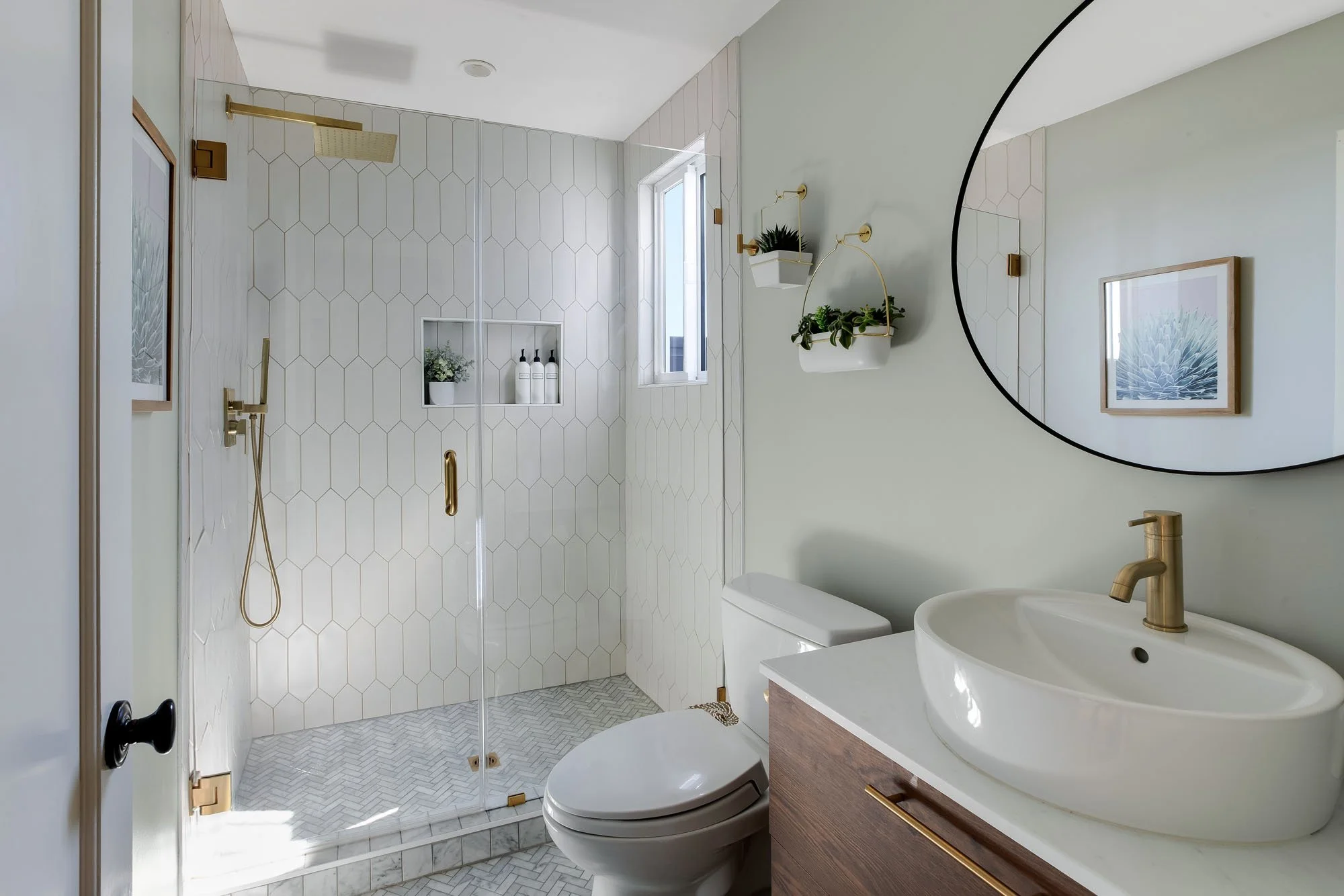 Fully renovated guest bathroom featuring a walk-in glass shower with white vertical picket tile, gold hardware, a wood vanity with a vessel sink, and a round black mirror.