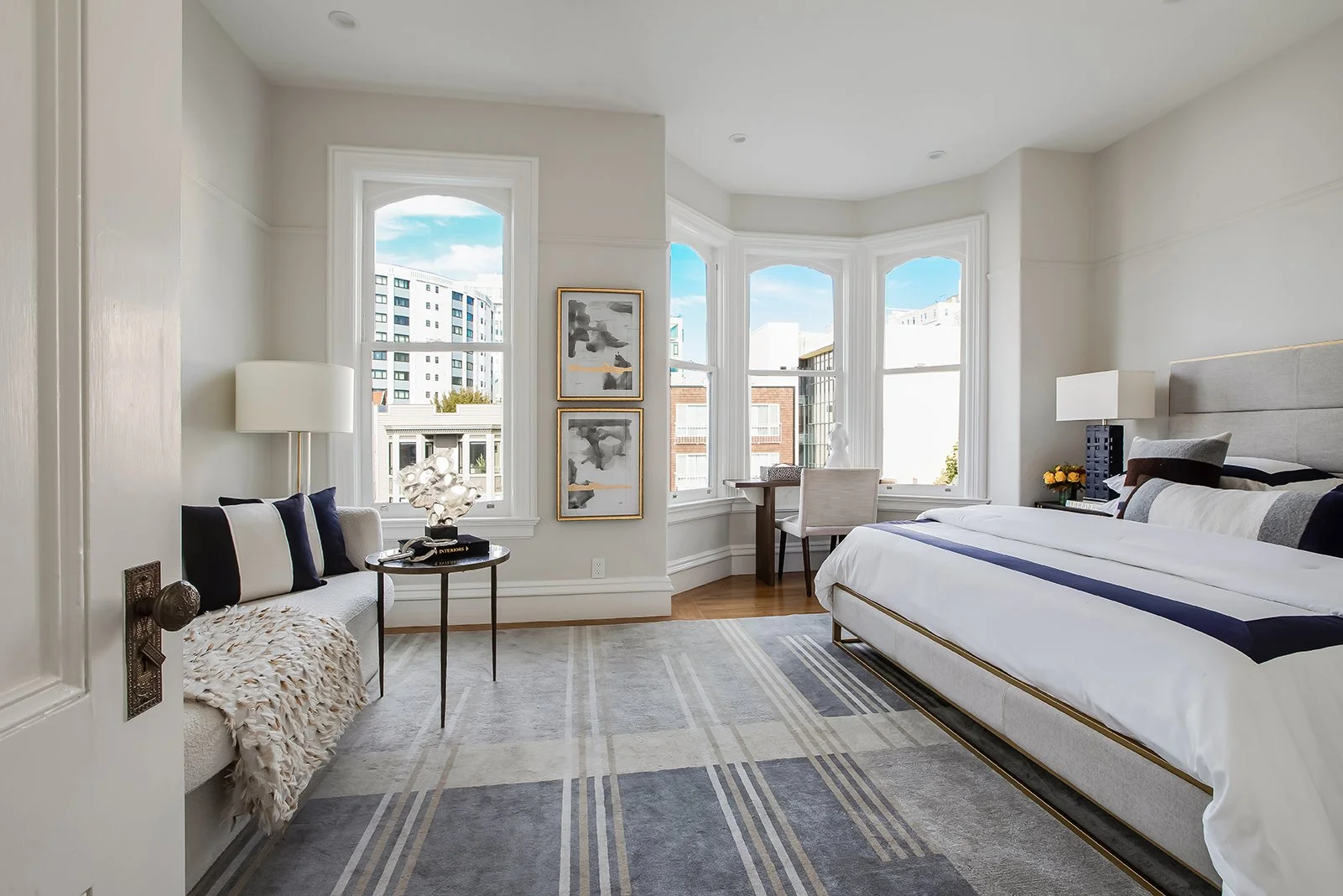 Expansive primary bedroom suite in a Pacific Heights Victorian featuring a signature curved bay window, dedicated workspace, and designer neutral decor.