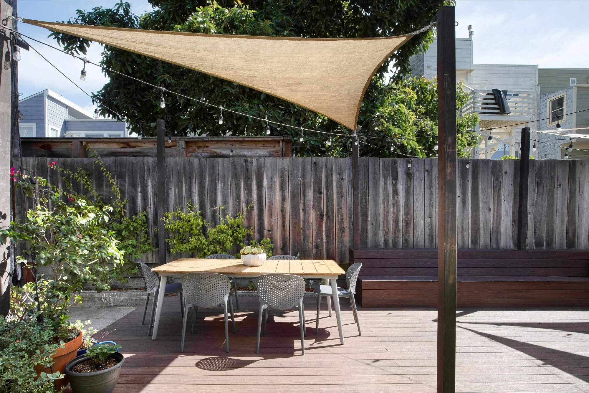 Rear yard dining area under a sail-shade canopy with string lights and garden plantings