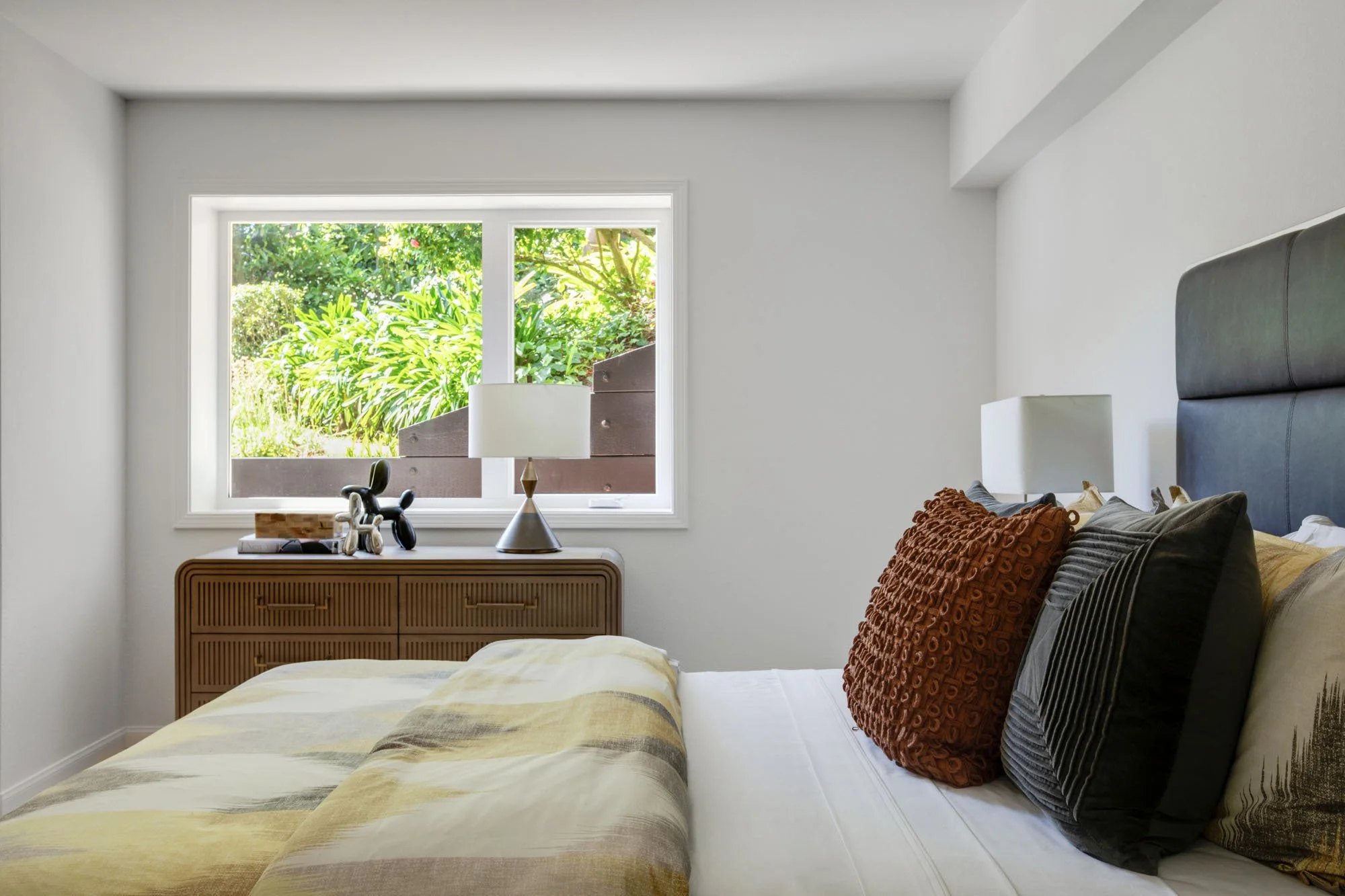 Close-up of a stylish bedroom interior highlighting a textured rust-colored throw pillow and a modern fluted dresser, with a clear view of a private, green San Francisco hillside through the window.