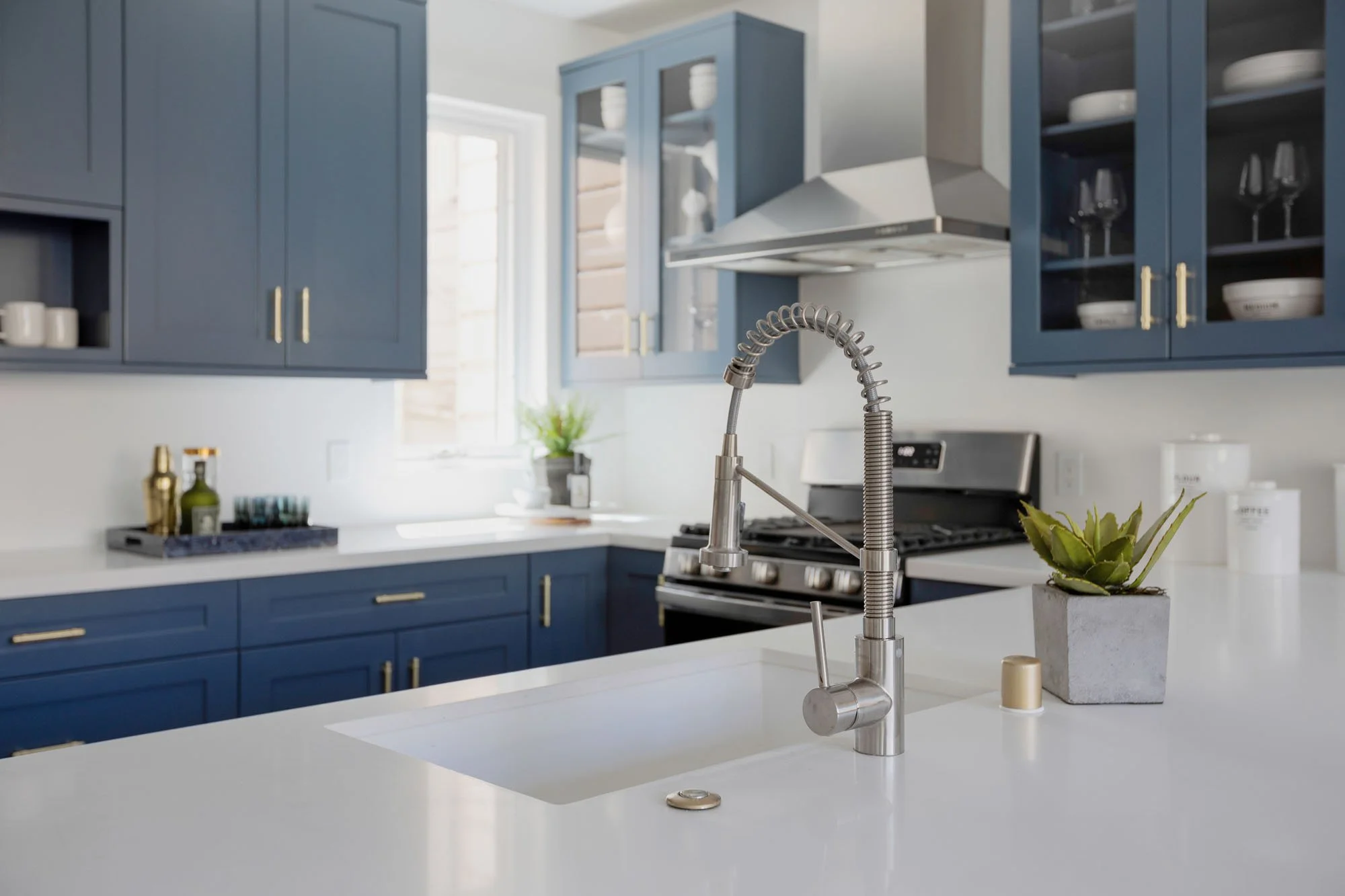 A close-up of a kitchen island featuring a white undermount sink and a professional-style stainless steel spring-neck faucet. The background shows blue cabinets, a stainless steel gas range, and a sleek vent hood.