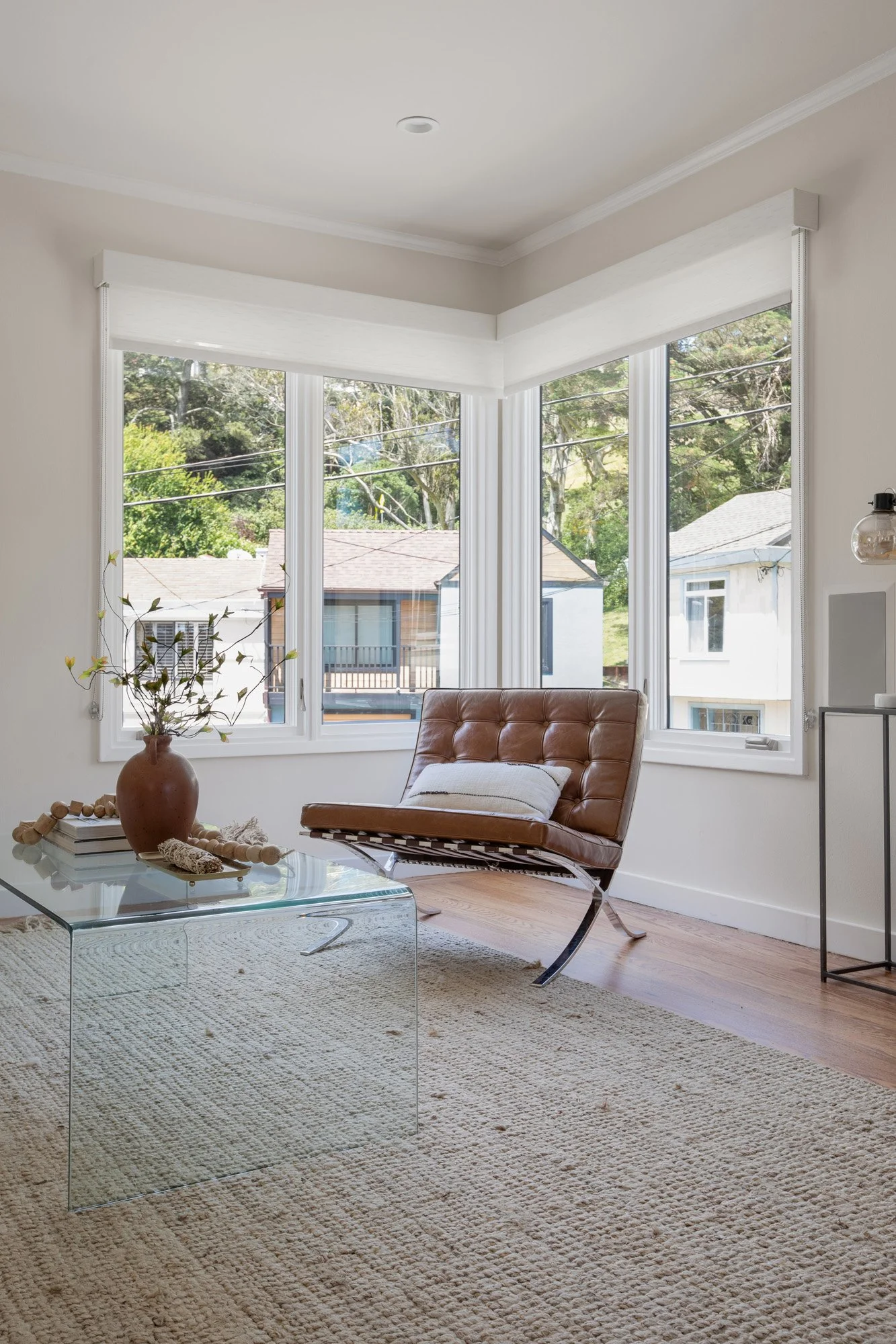 Minimalist living room corner with corner windows, a Barcelona-style leather chair, and a clear glass waterfall coffee table.