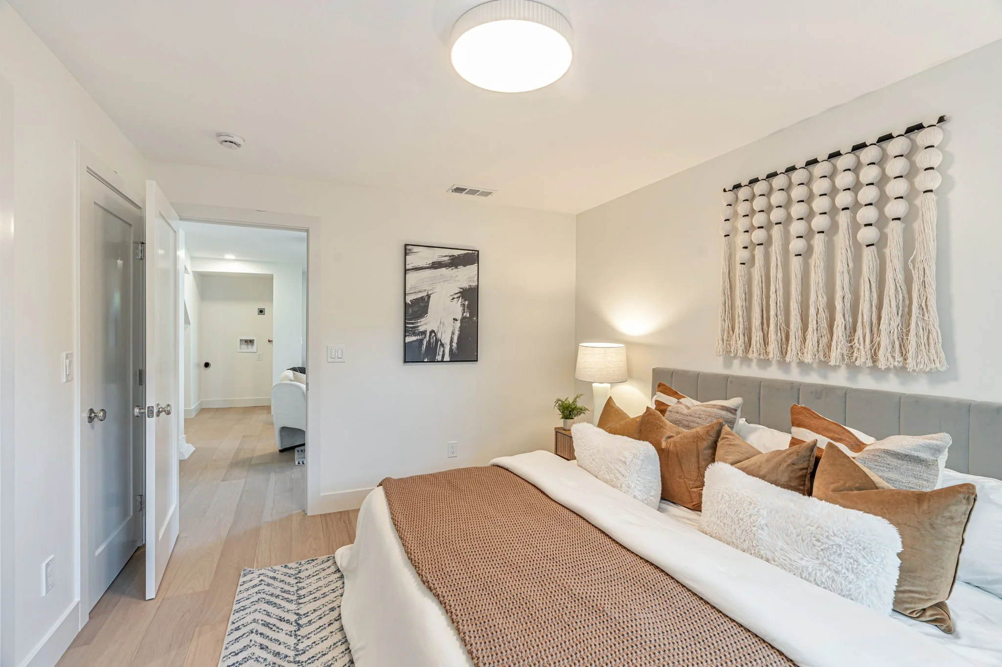 Boho-modern guest bedroom suite with a large decorative macrame wall hanging, rust-colored textured pillows, and a view into the lower-level media room.