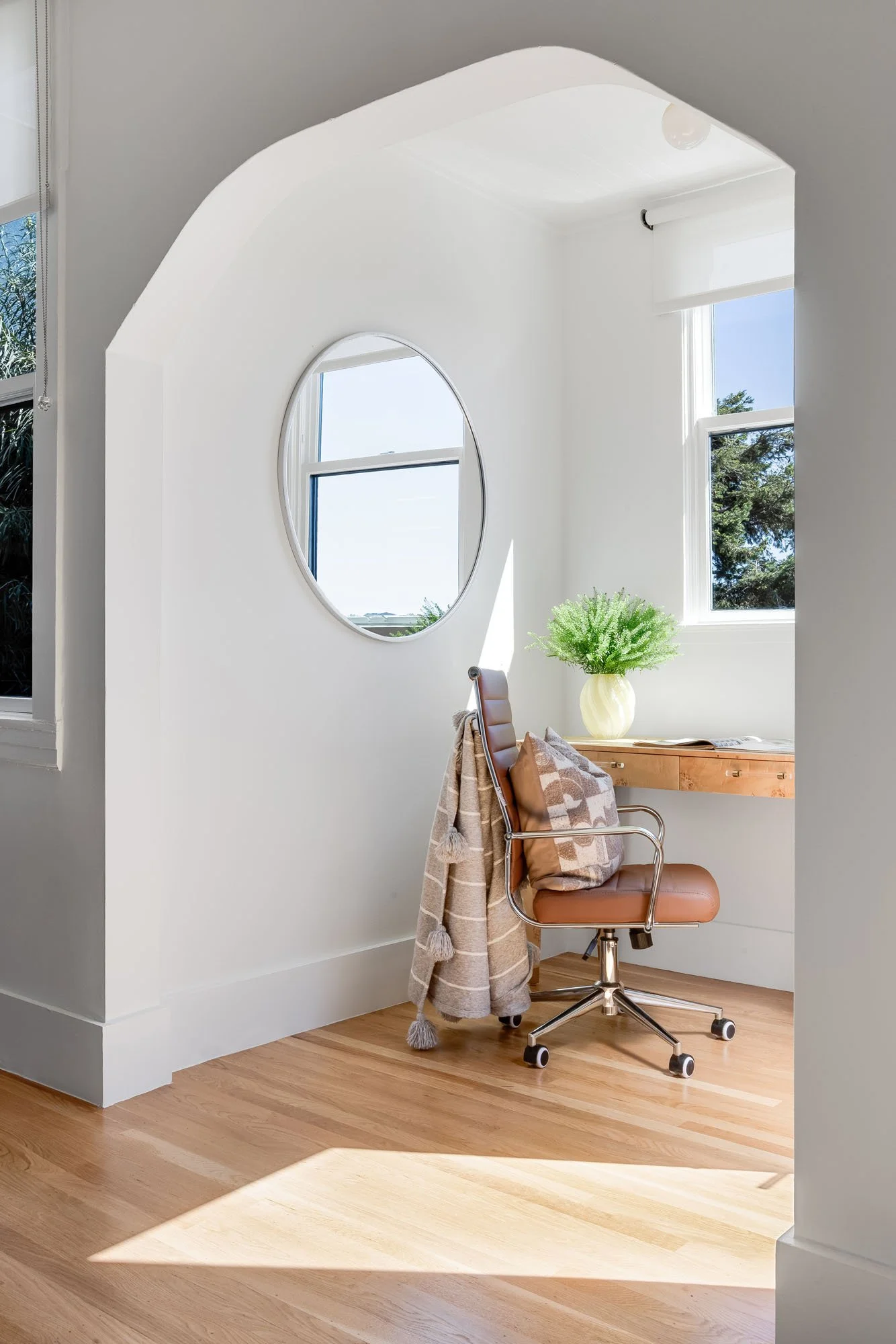 Close-up of a cozy home office nook framed by a classic architectural arch, featuring a wooden desk, a leather office chair, and a large circular mirror.