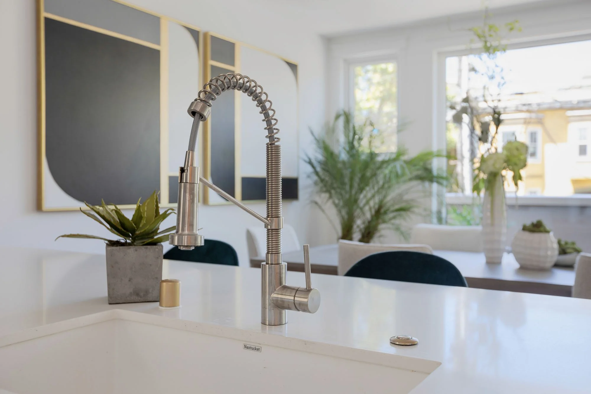 Close-up of a commercial-style stainless steel spring-neck kitchen faucet on a white quartz countertop with a view of the modern dining area in the background.