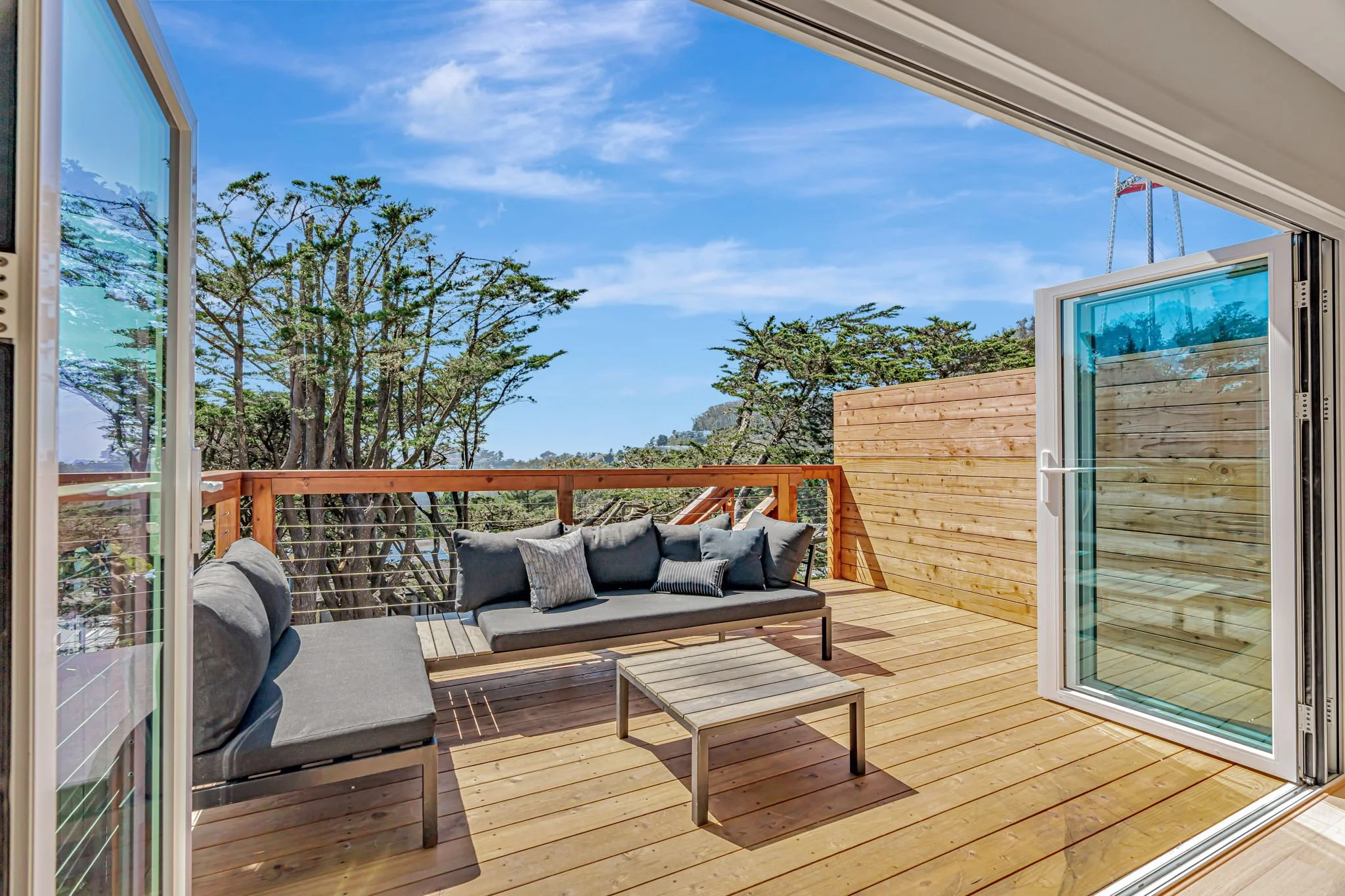 A view from inside looking out through wide-open accordion doors onto a spacious wooden deck. The deck is furnished with grey modern outdoor sofas and a coffee table, overlooking a canopy of evergreen trees.