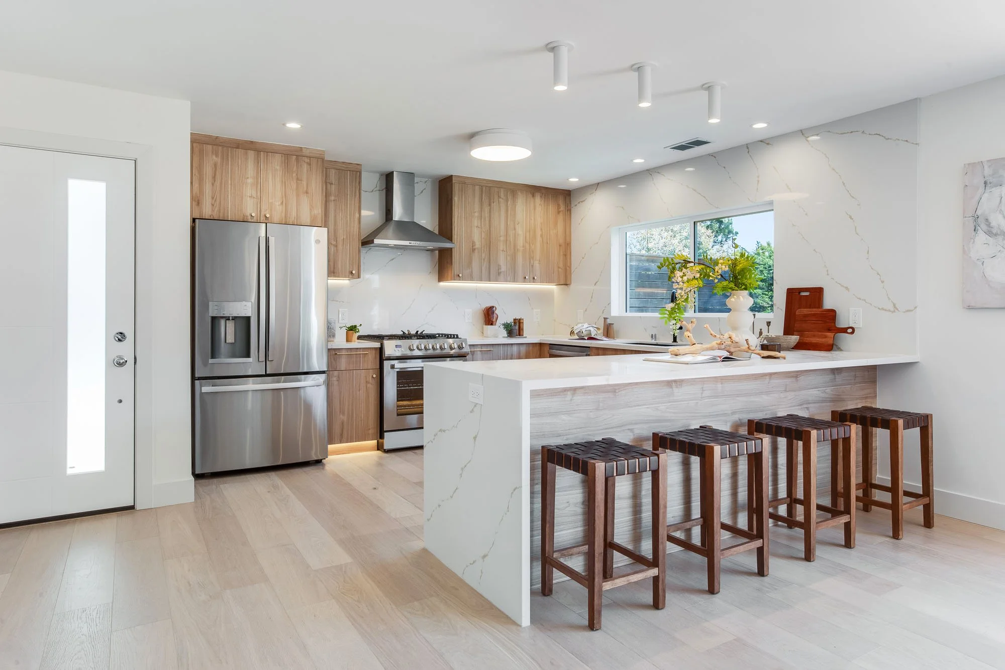 A wide, bright view of the modern kitchen featuring a large waterfall-edge marble-look quartz island. Four dark wood stools with woven leather seats are tucked under the breakfast bar, while the background shows light wood cabinetry and high-end stai