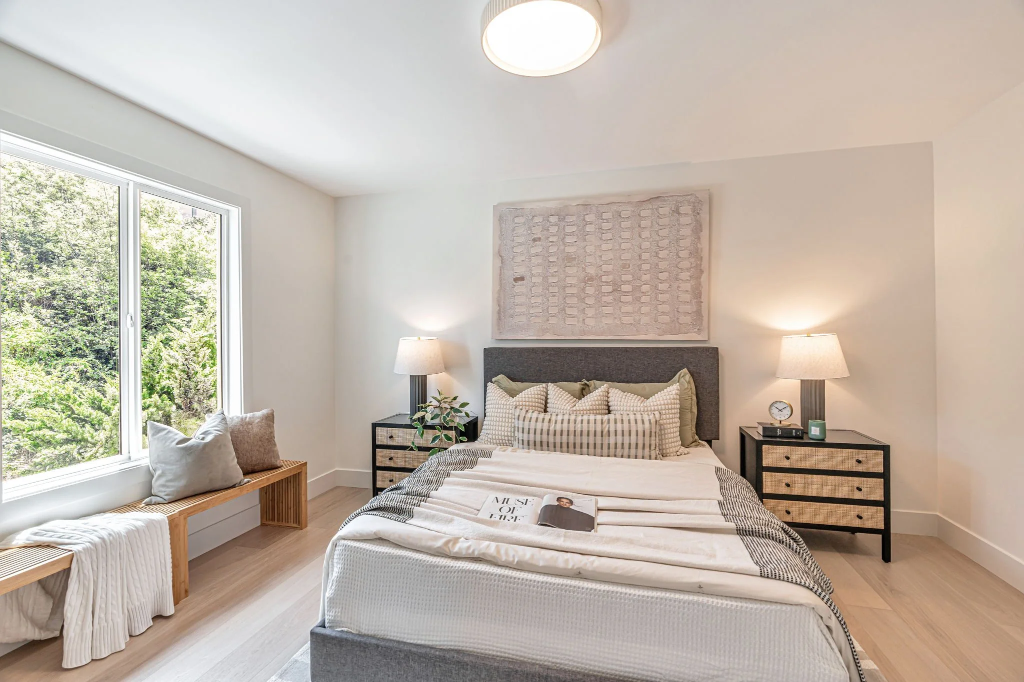 A serene primary bedroom with light hardwood floors and a large window overlooking a lush green canopy. The room features a grey upholstered headboard, black nightstands with woven cane drawers, and a long wooden bench under the window.