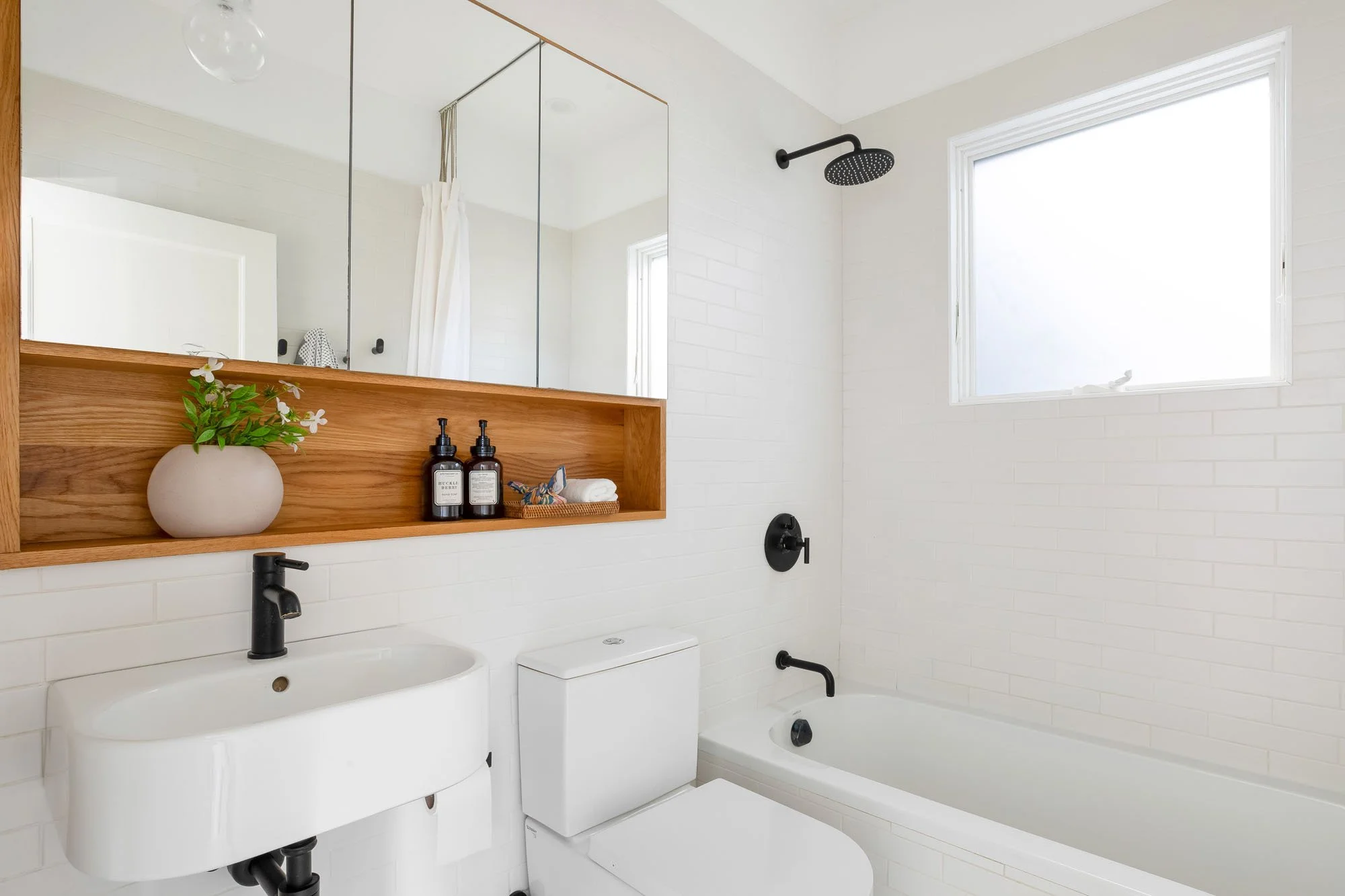 A modern, bright bathroom with white subway tile, a floating sink, a bathtub-shower combo with matte black fixtures, and a custom wood-framed medicine cabinet.